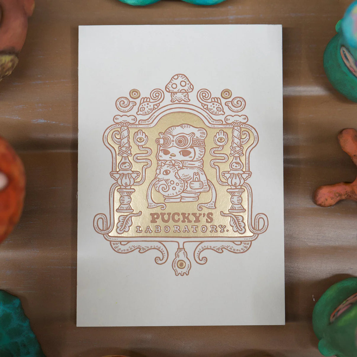 Notebook Laboratory - Preorder: An ornate art card featuring a cat-scientist in goggles mixing potions, titled PUCKY'S LABORATORY, surrounded by colored clay on a wooden table.