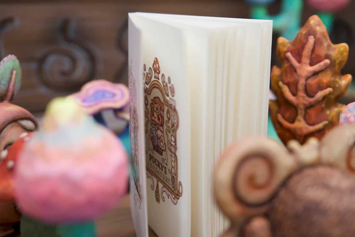 Notebook Laboratory - Preorder: An open zine featuring a cartoon character with goggles, surrounded by whimsical, handmade figurines like mushroom caps and spirals, evoking playful creativity.