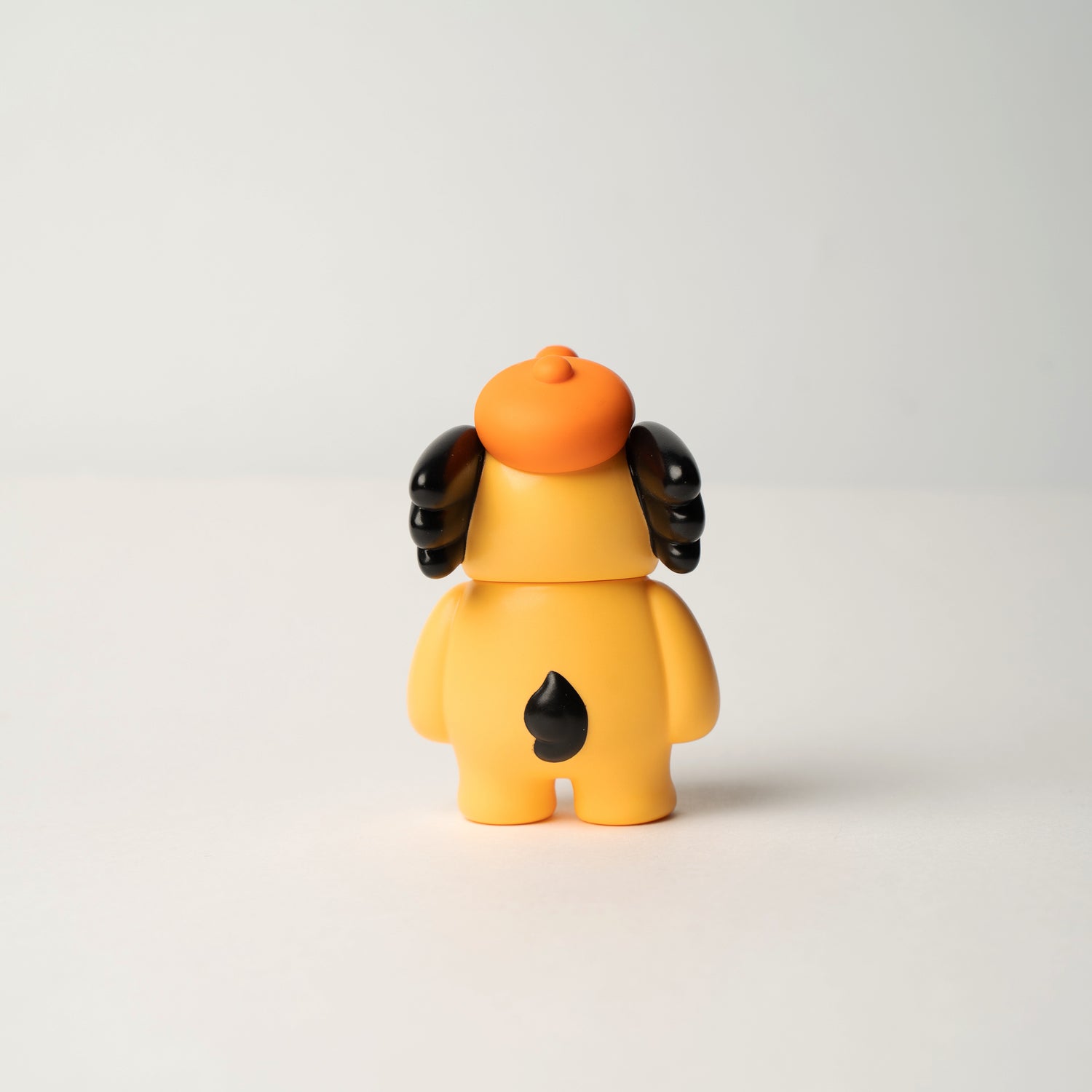 Yellow vinyl toy figure, Elfie 100% OHIGEnoPON Edition, with floppy black ears and an orange beret, displayed from the back on a white surface.