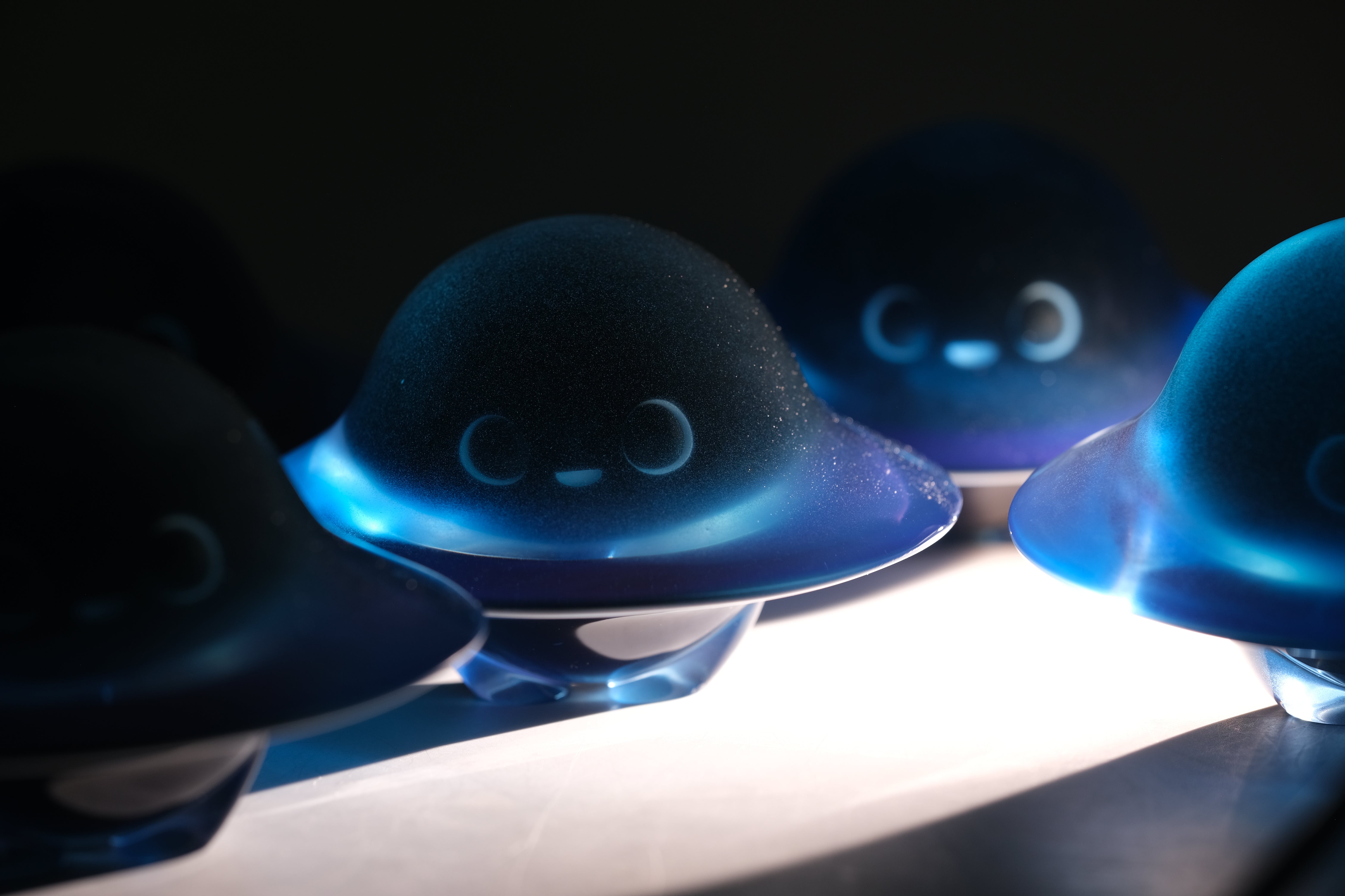 Glow in the Darkmite! by Yutoos features dome-shaped, translucent resin figurines with smiling faces, giving a bioluminescent effect under focused lighting.