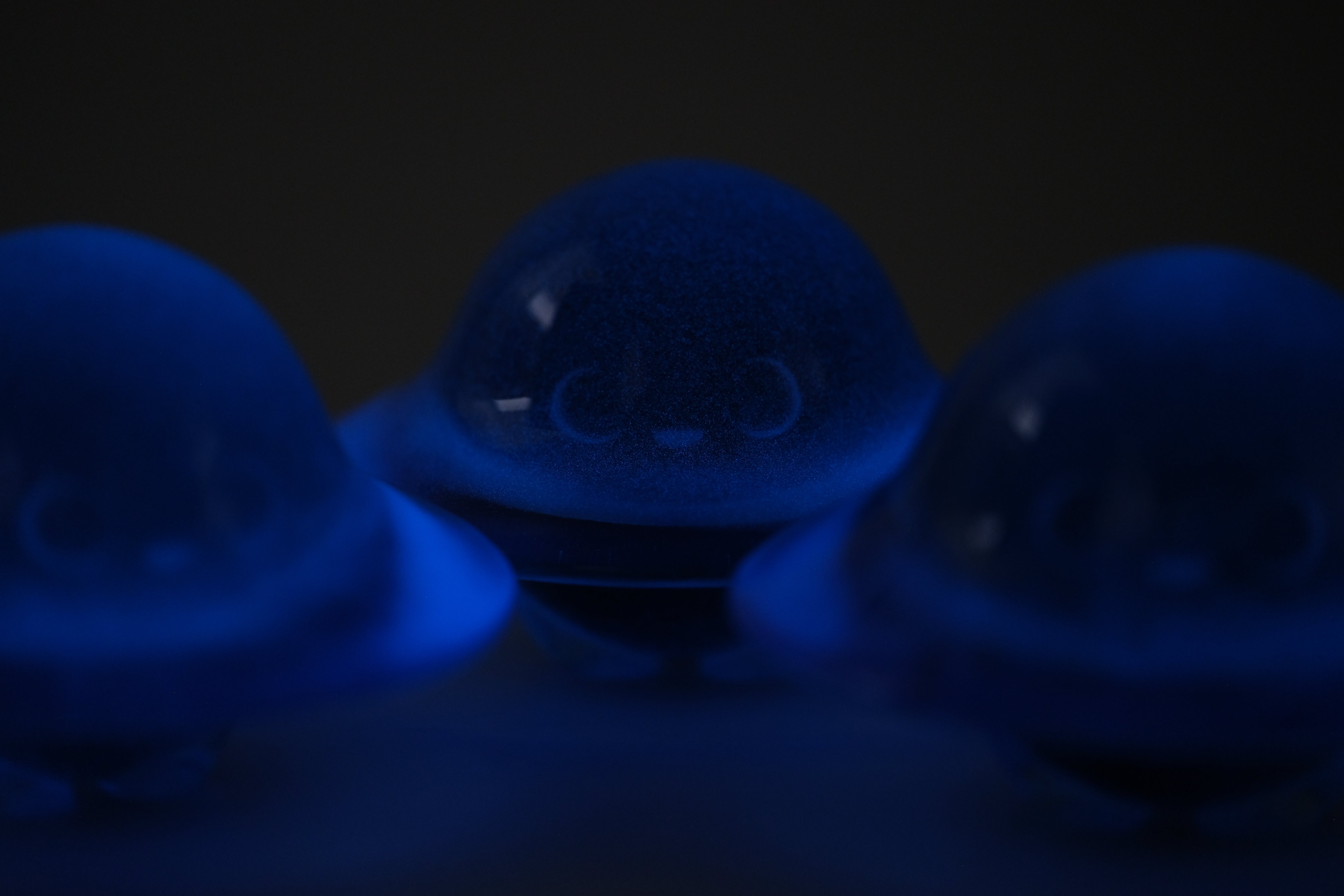 Glow in the Darkmite! by Yutoos: Three small, translucent blue resin figures with smiling faces, the center one sharply focused against a dark background.