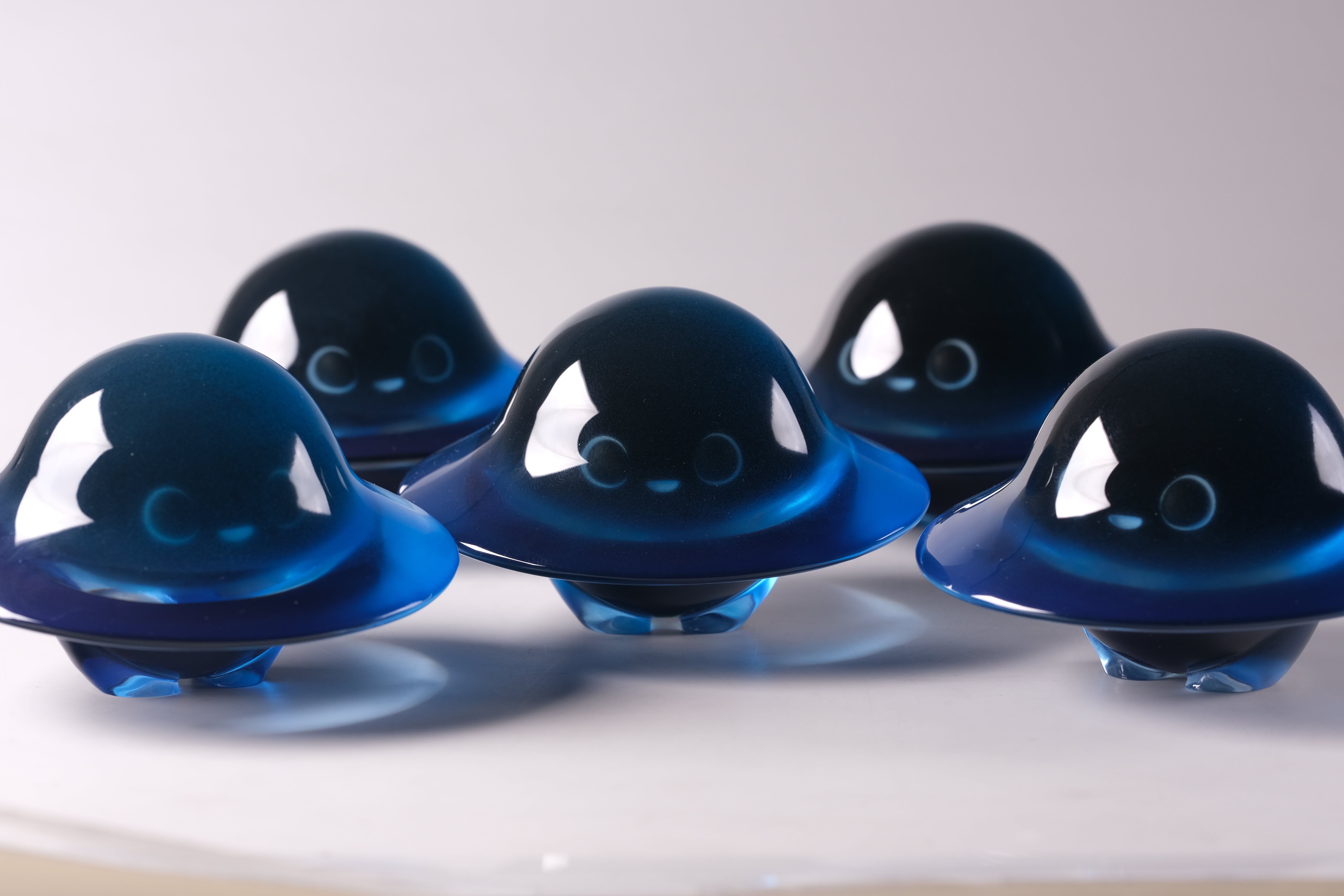 Glow in the Darkmite! by Yutoos features five glossy, deep-blue, translucent resin figurines with cartoon faces, resembling flying saucers, highlighting their shiny, jewel-like finish.