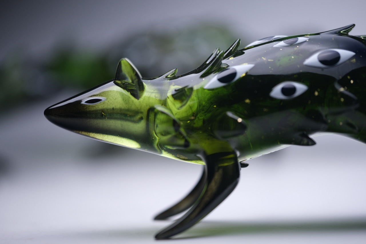 Close-up of Bogwalker Oracle by Faunwood, a hand-blown green glass sculpture resembling a stylized fish with fin-like protrusions and painted eye motifs.