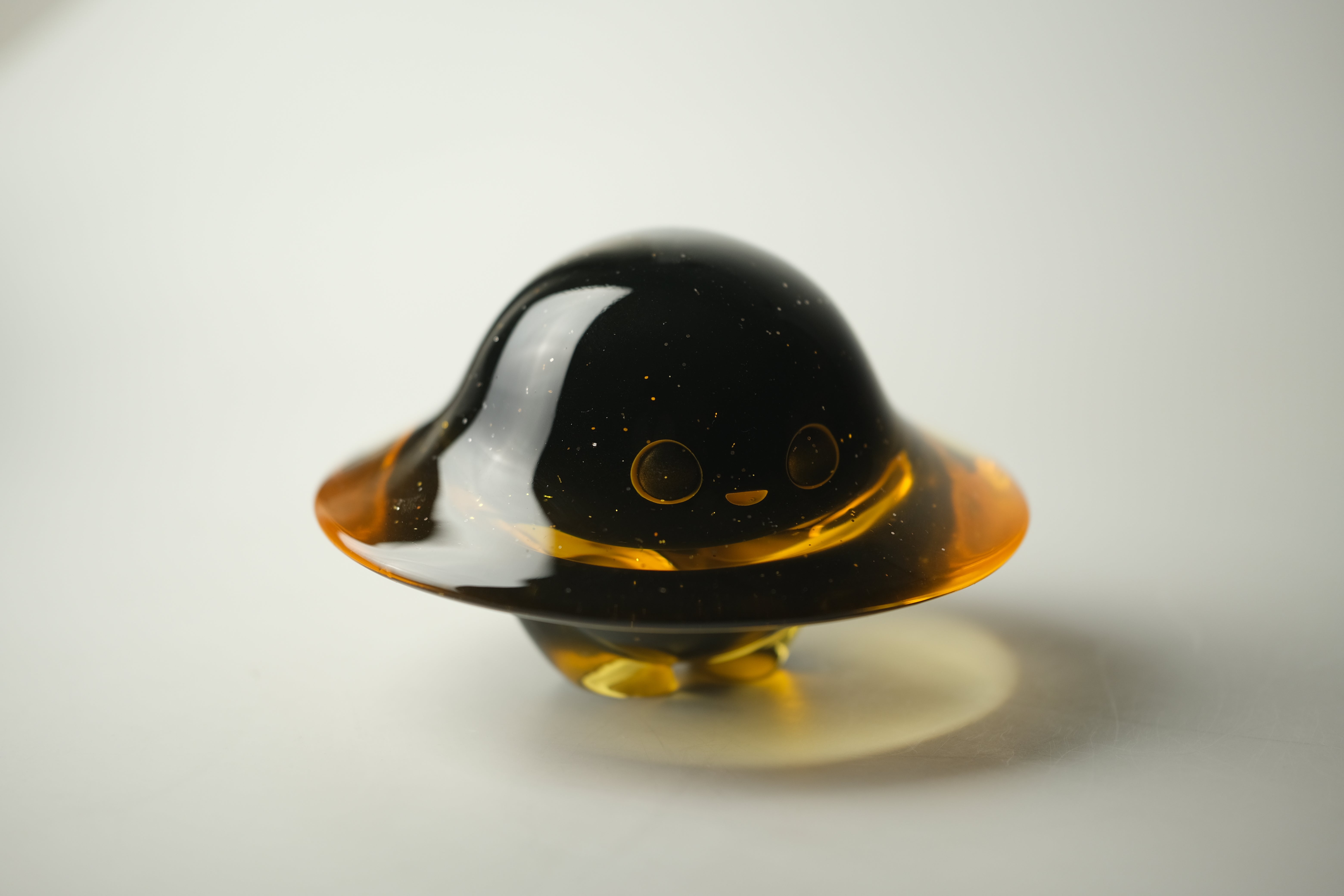 Darkmite by Yutoos: A glossy black resin figurine resembling a UFO, featuring gold flecks, an amber rim, and a smiling face. Edition of 50, each unique.
