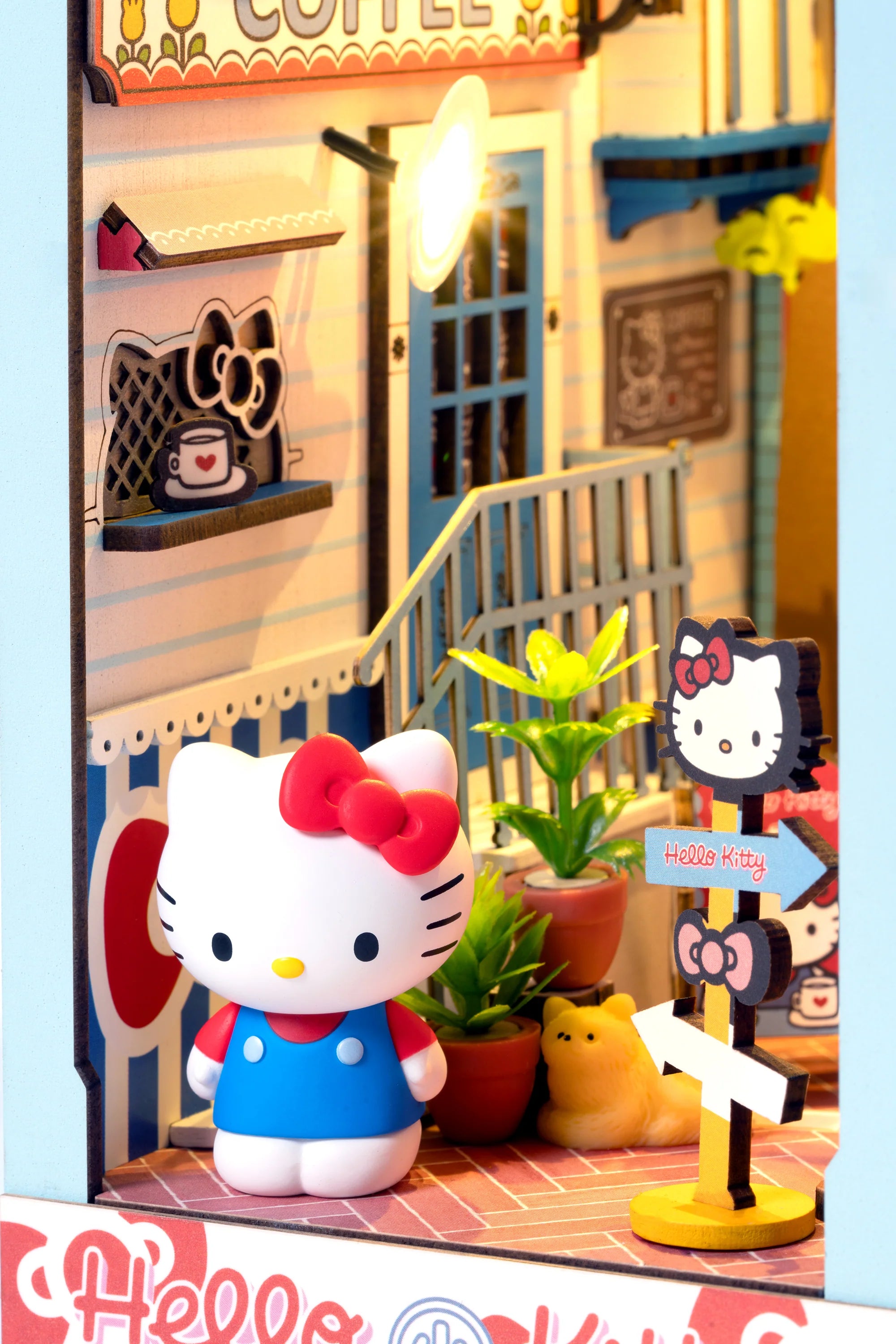Hello Kitty® Town Square Booknook diorama with Hello Kitty figurine, coffee shop façade, potted plants, directional signs, and touch-activated LED lights for a cozy display.
