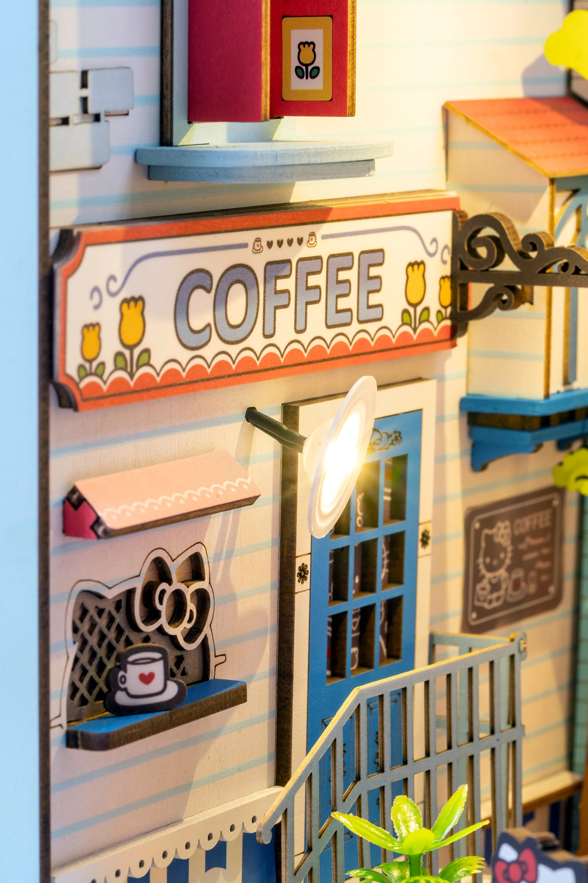 Hello Kitty® Town Square Booknook: Miniature diorama with COFFEE sign, tulip trim, blue door, menu, plant, and Hello Kitty illustration, featuring touch-activated LED lights.