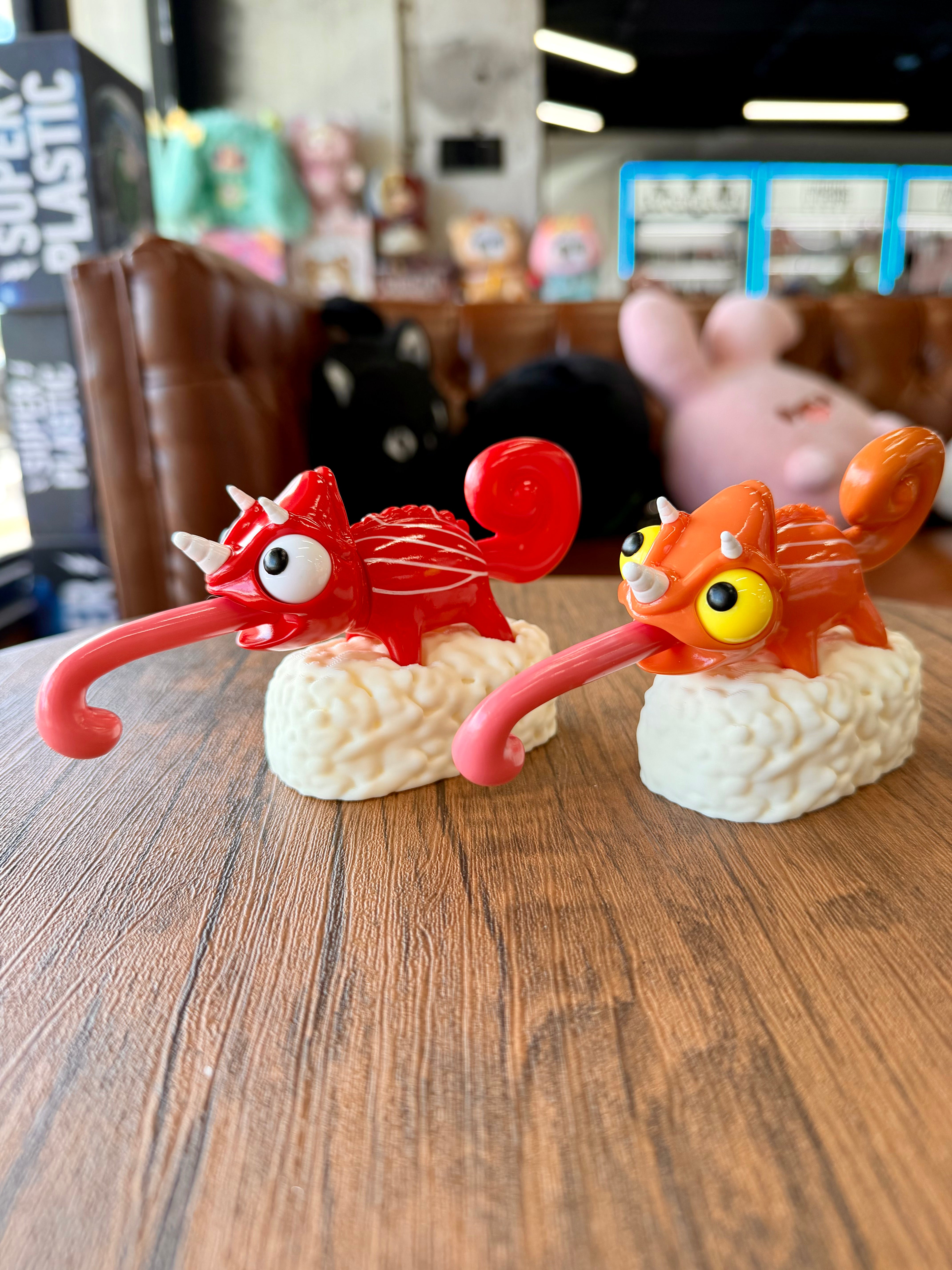 Two cartoonish chameleon figurines, Sushi Gogo Chameleons, perched on sushi rice mounds, with curled tongues and horn-like protrusions, displayed on a wood-grain tabletop.