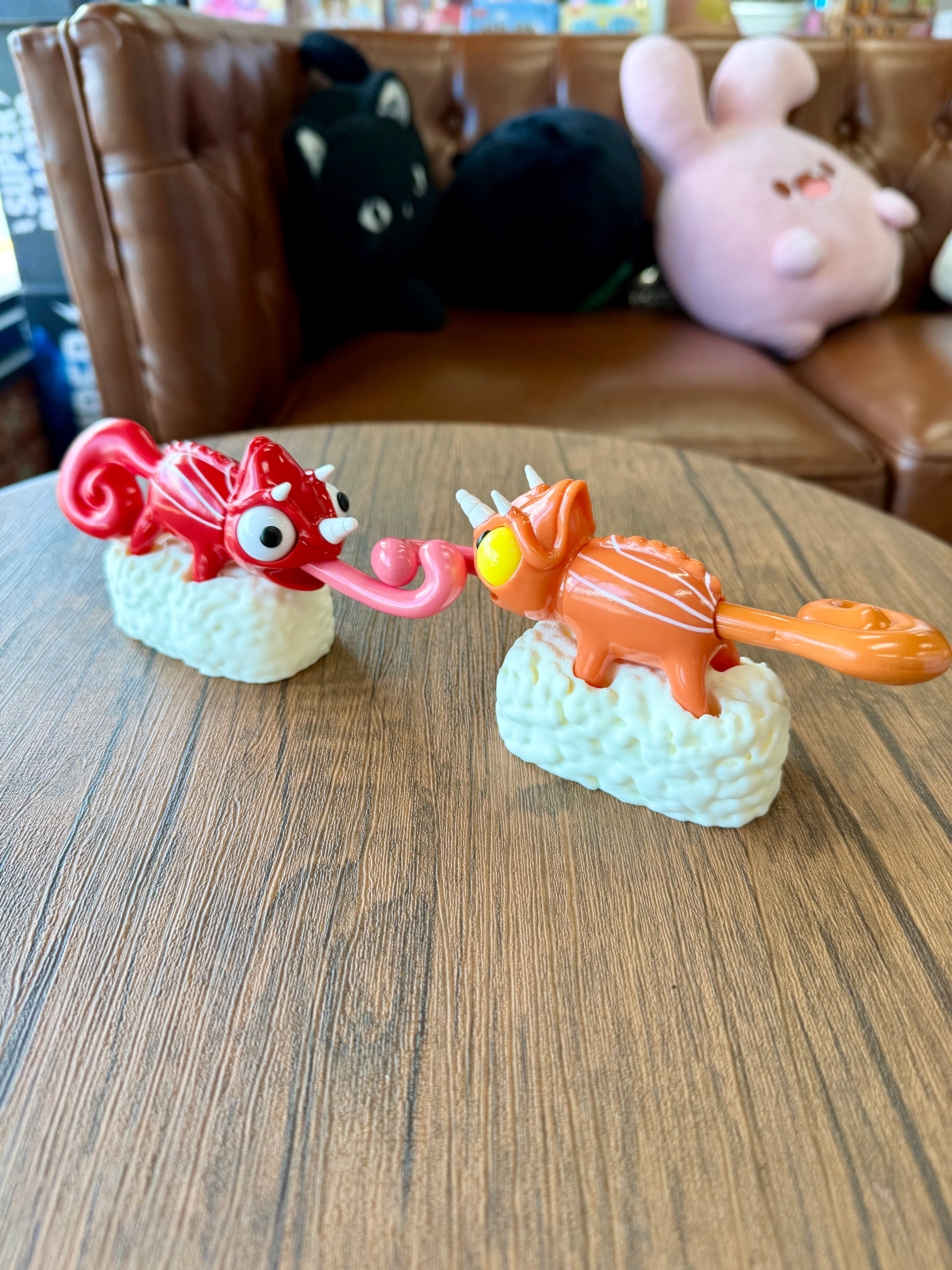 Sushi Gogo Chameleon vinyl figurines, one red with horns and tongue, the other orange with stripes, perched on sushi rice bases, in a playful setting.