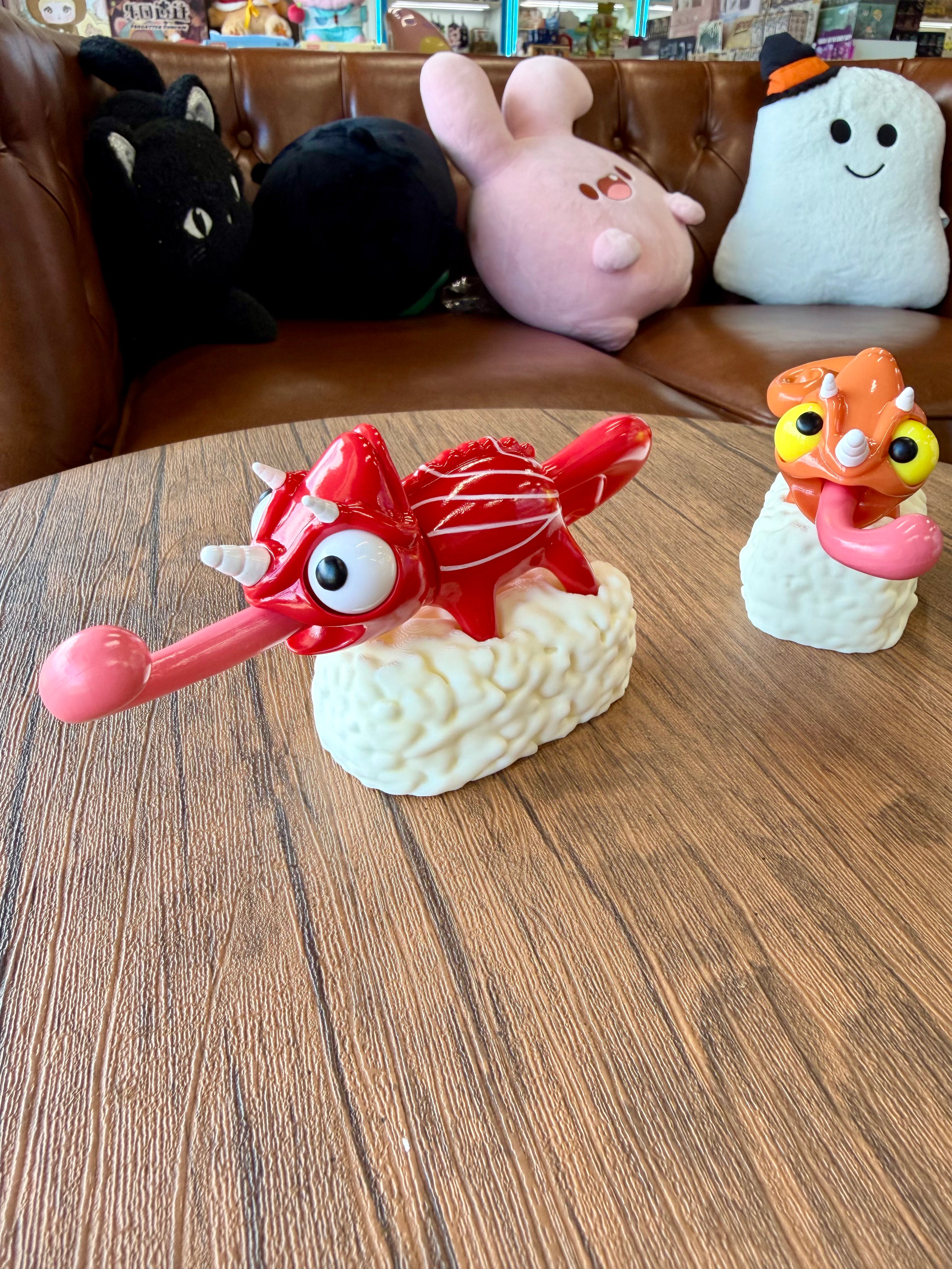 Sushi Gogo Chameleon vinyl figures, 15cm, depicting chameleon-creatures on sushi, with cartoonish eyes and horns, set on a wooden table by a leather sofa.