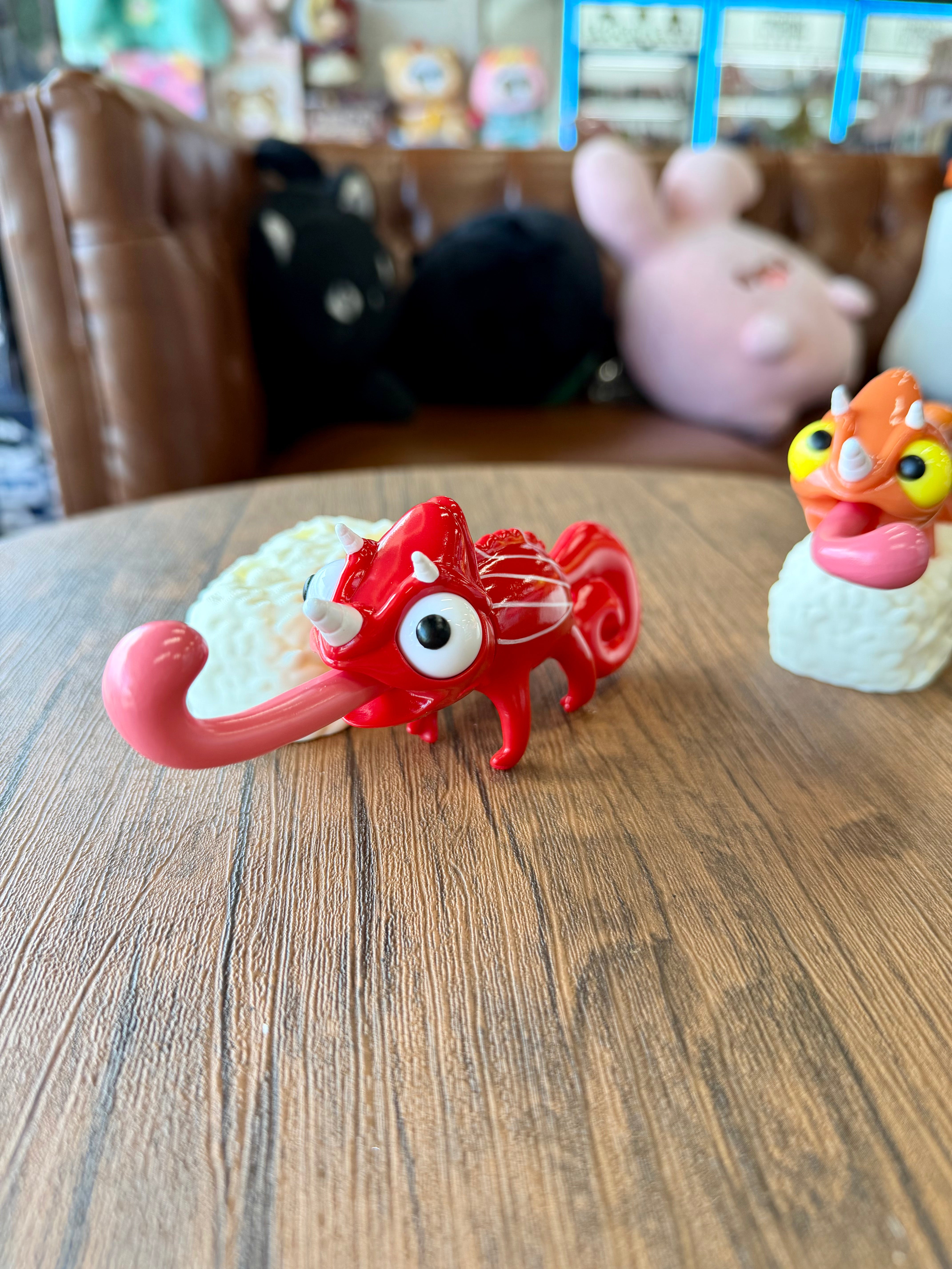 Sushi Gogo Chameleon: A bright red, stylized toy with big eyes, white horns, and a long pink tongue, sitting on a table.
