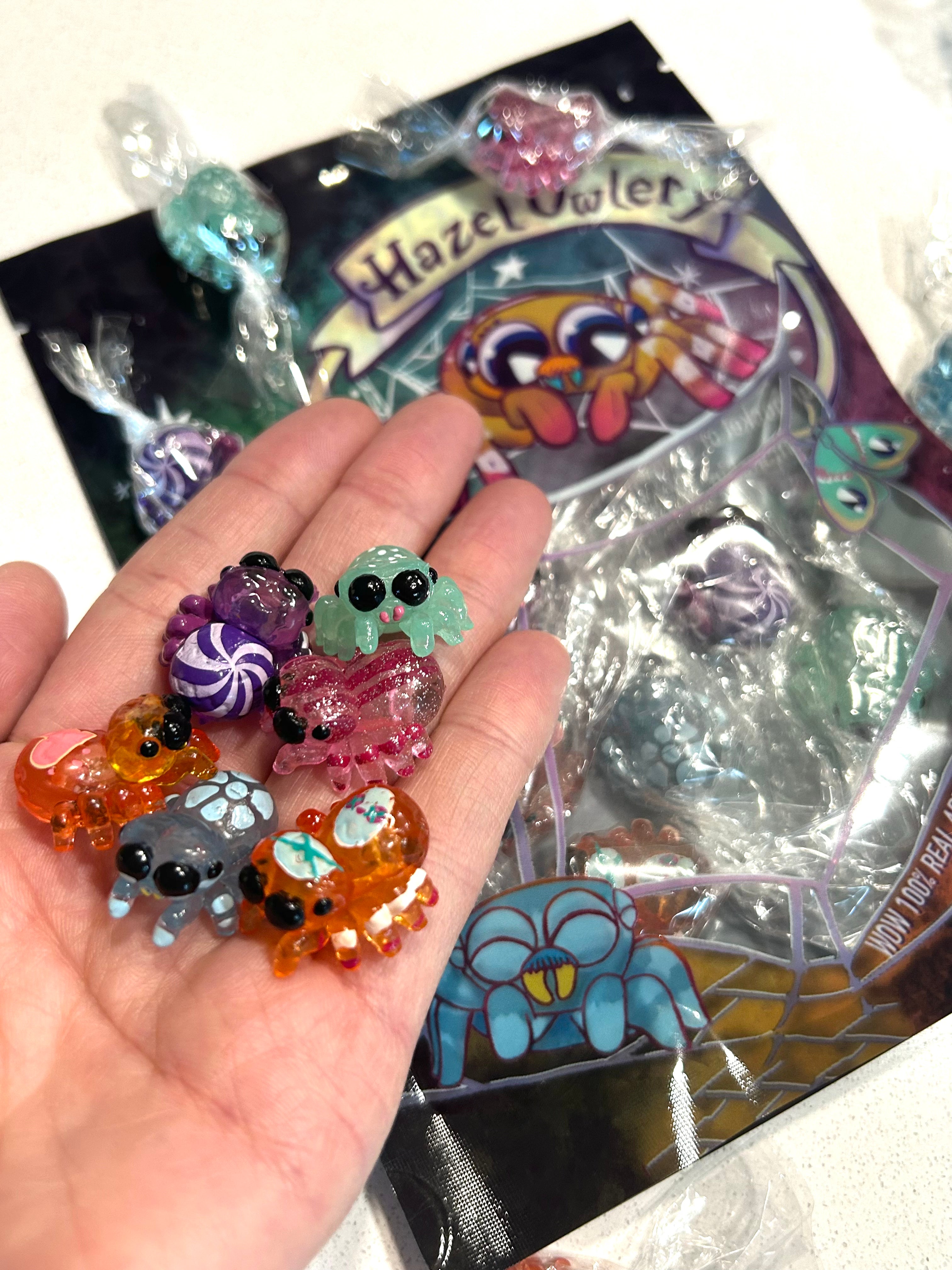 A hand holding small plastic spiders, a toy, and a candy from Spidey Sweets by Hazel Owlery.