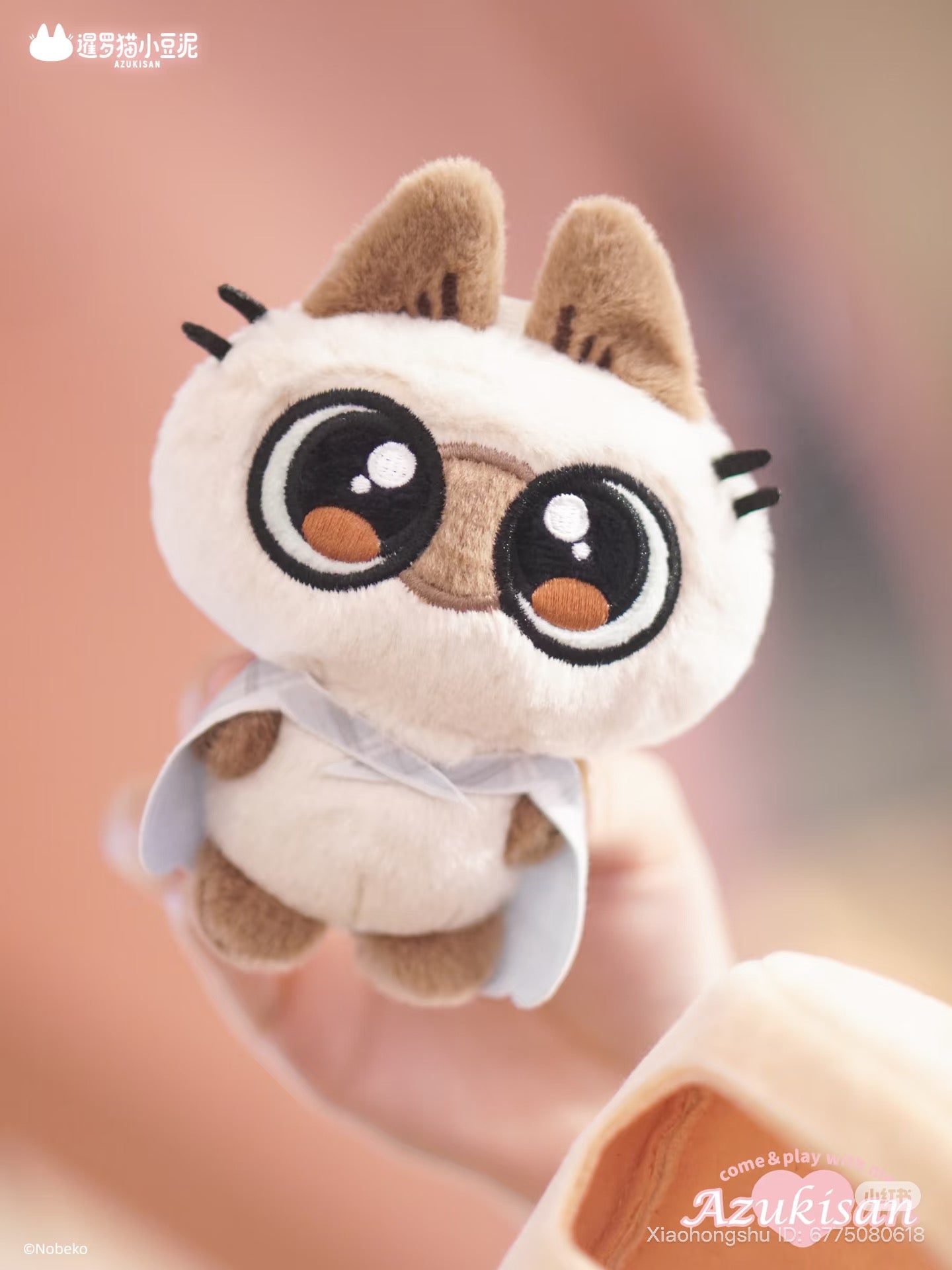 Azukisan's Daily Life Series plush pendant, a stylized cat toy with embroidered eyes and a cape, held up against a blurred background. Preorder available.