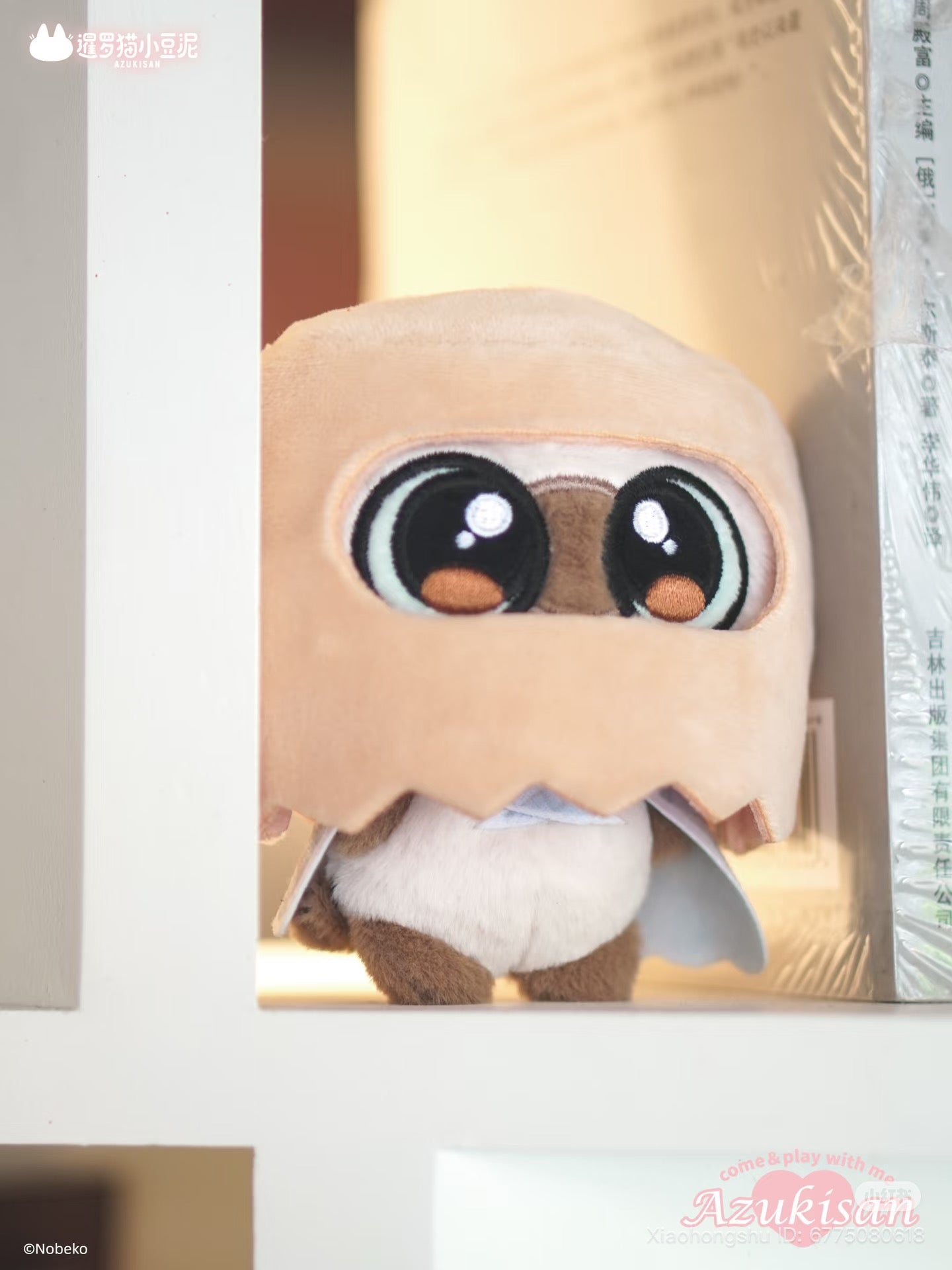 Azukisan's Daily Life Series plush pendant blind box, featuring a plush toy with round eyes, tiny wings, and a paper-bag hood, displayed on a shelf with books.