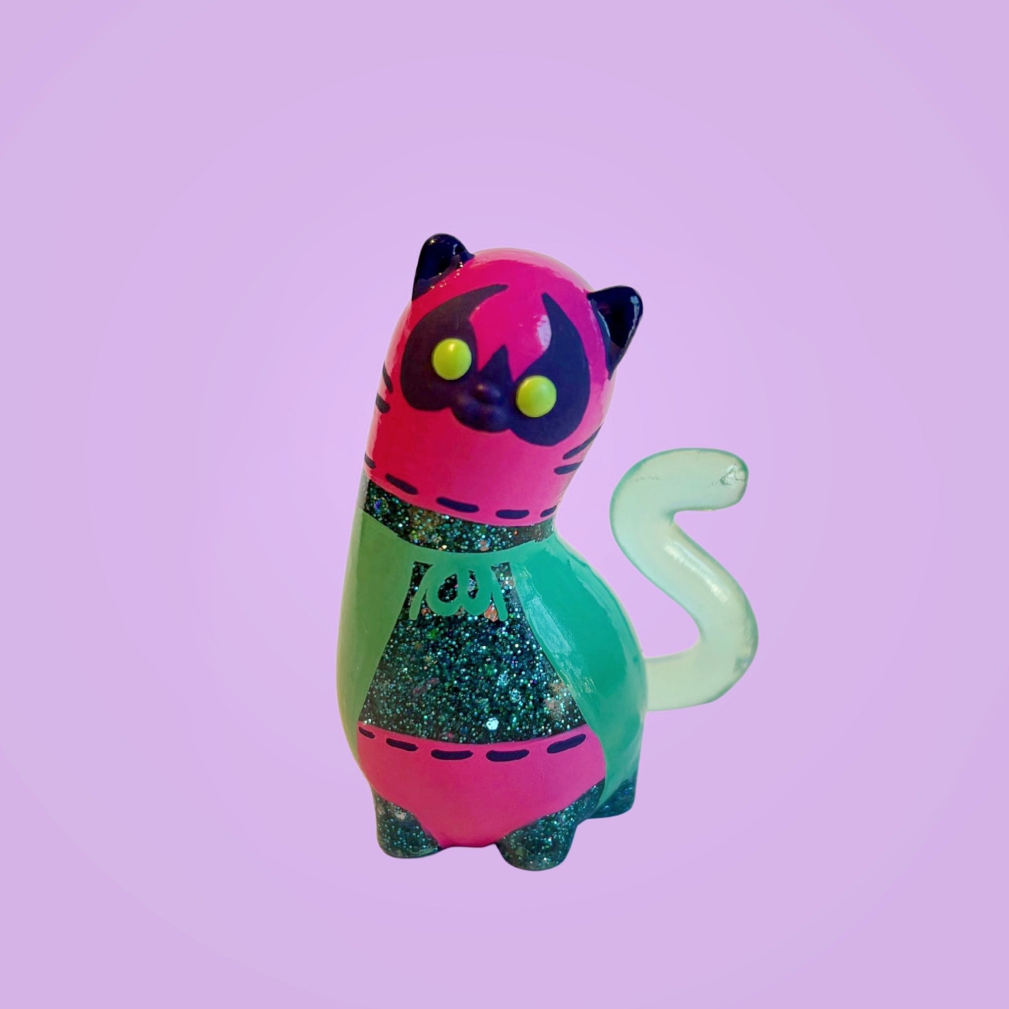 Spencer by Ghost Fox Toys: A whimsical, resin cat figurine with a bright pink head, large green eyes, and a glittery green body, showcasing a playful, minimalist design.