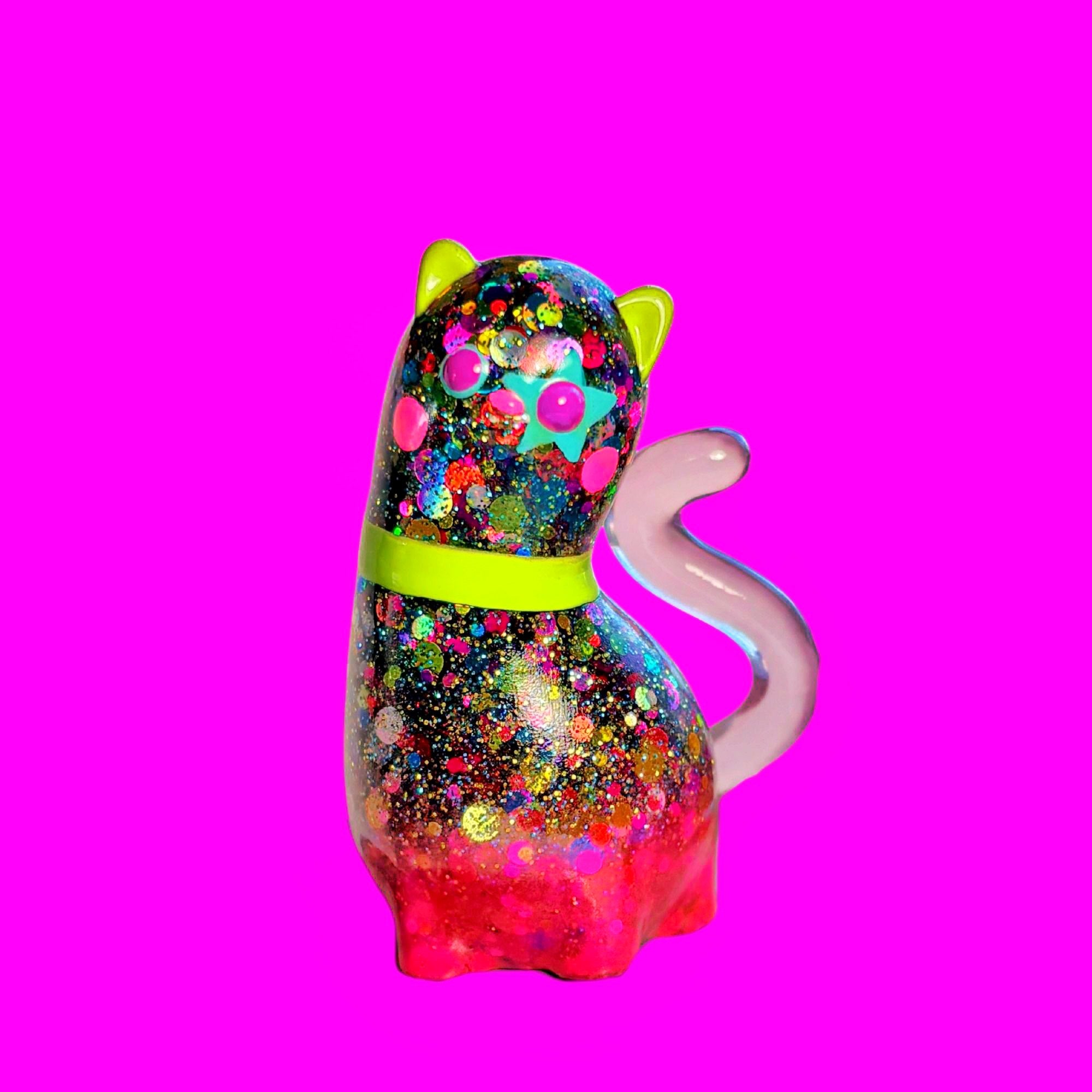 Spencer by Ghost Fox Toys: A small, glittery, resin cat figurine with neon ears, star-shaped eye, and pastel-pink tail, set against a vibrant backdrop.