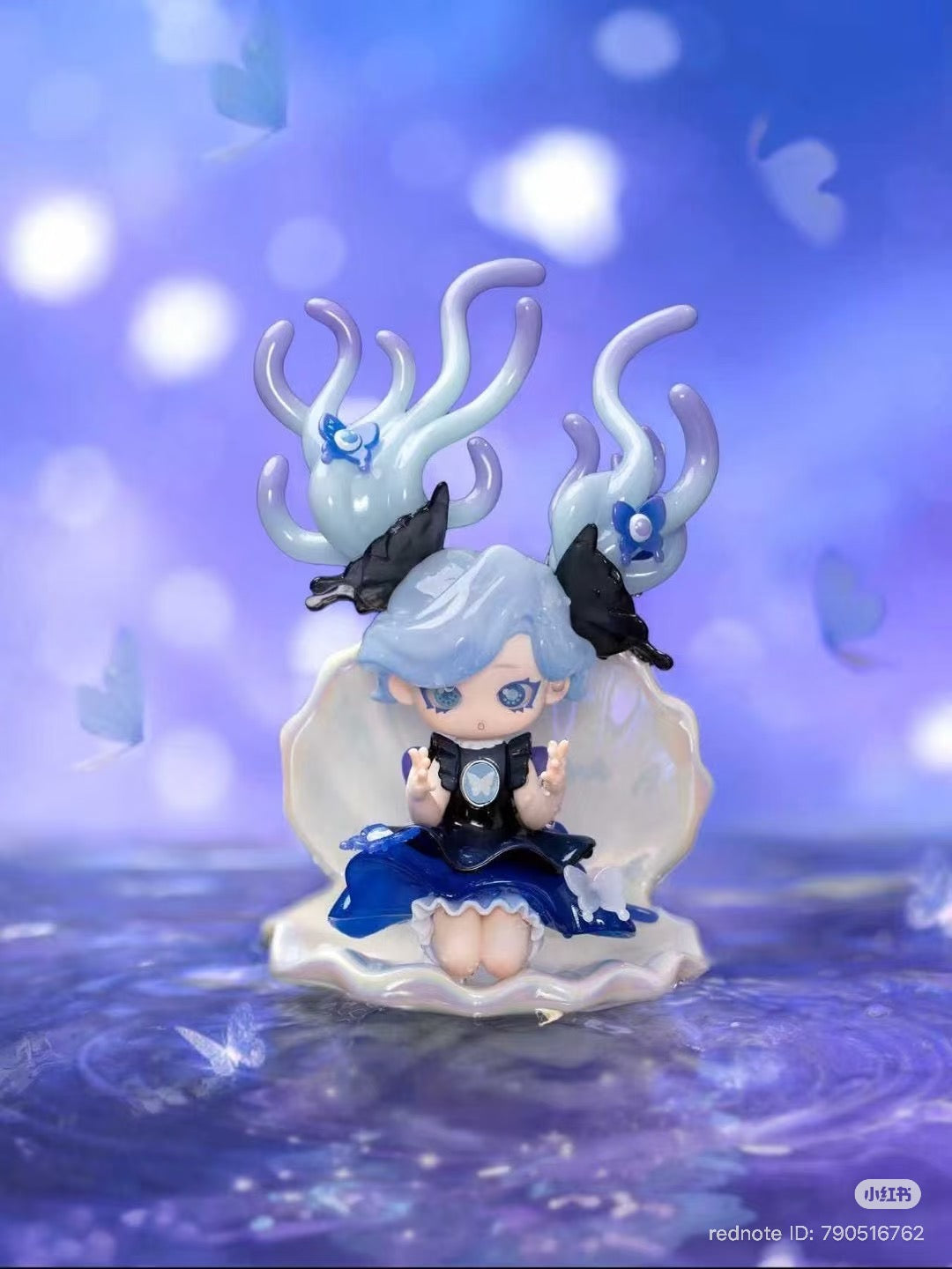 Chibi-style figurine from Memimo-Unusual Dream Blind Box Series, featuring pastel-blue hair and dark blue dress, seated in a clamshell with glowing bokeh accents.