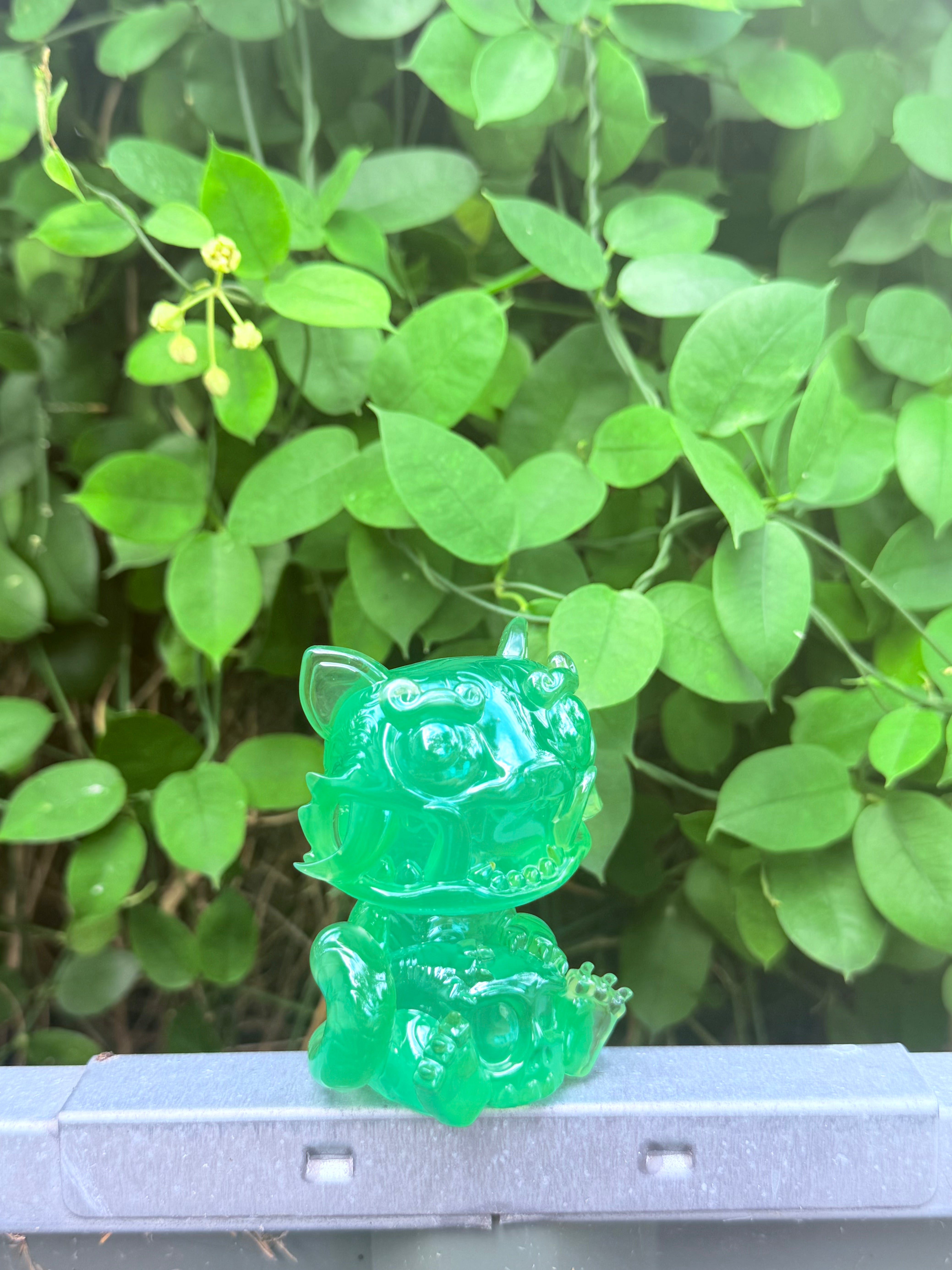 RuRu by Tomodachi Island, a small, translucent green monster-cat resin figurine, sits on a metal ledge with lush green leaves in the background.