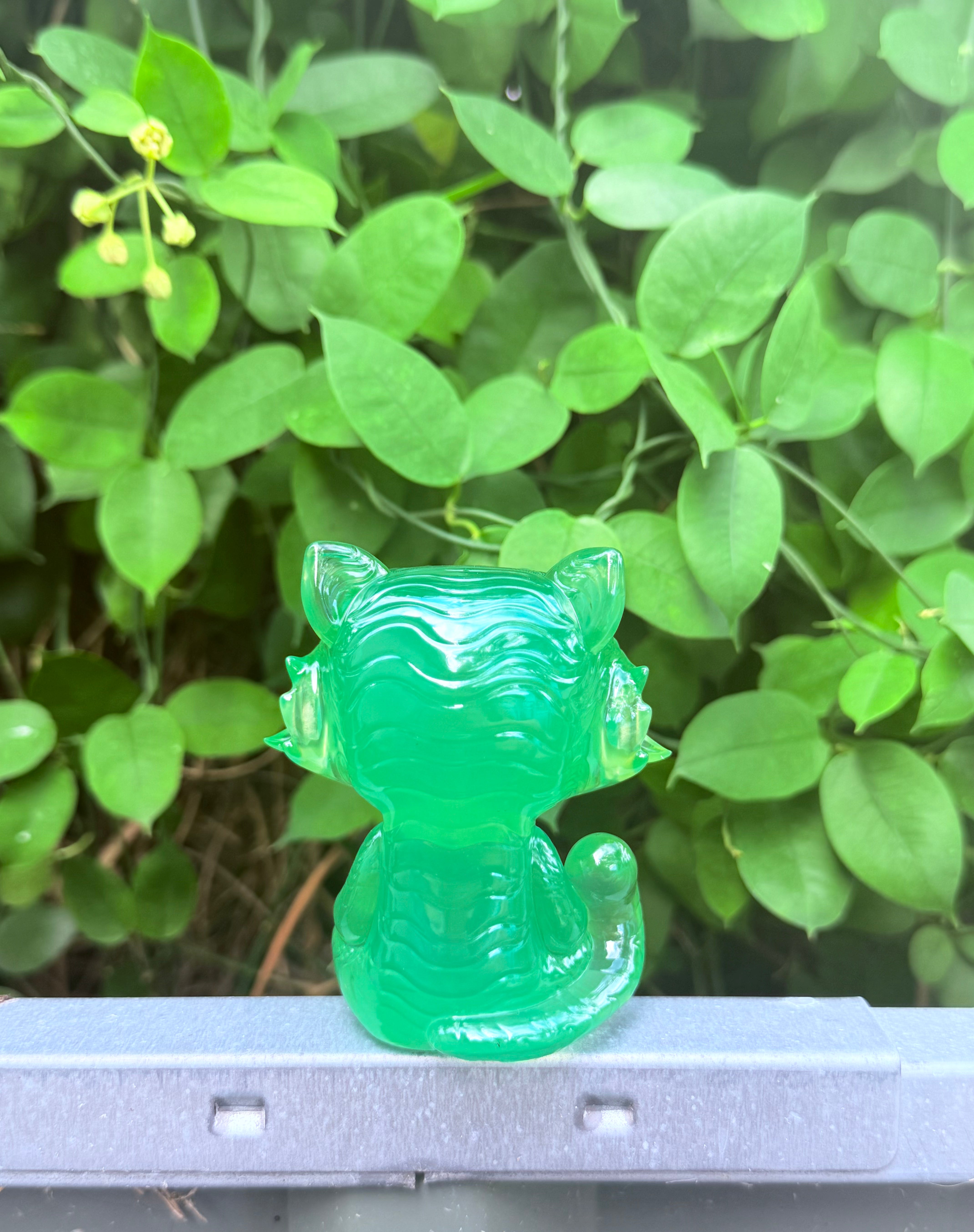 RuRu by Tomodachi Island, a small translucent resin cat figurine, perched on a grey surface, surrounded by green foliage.