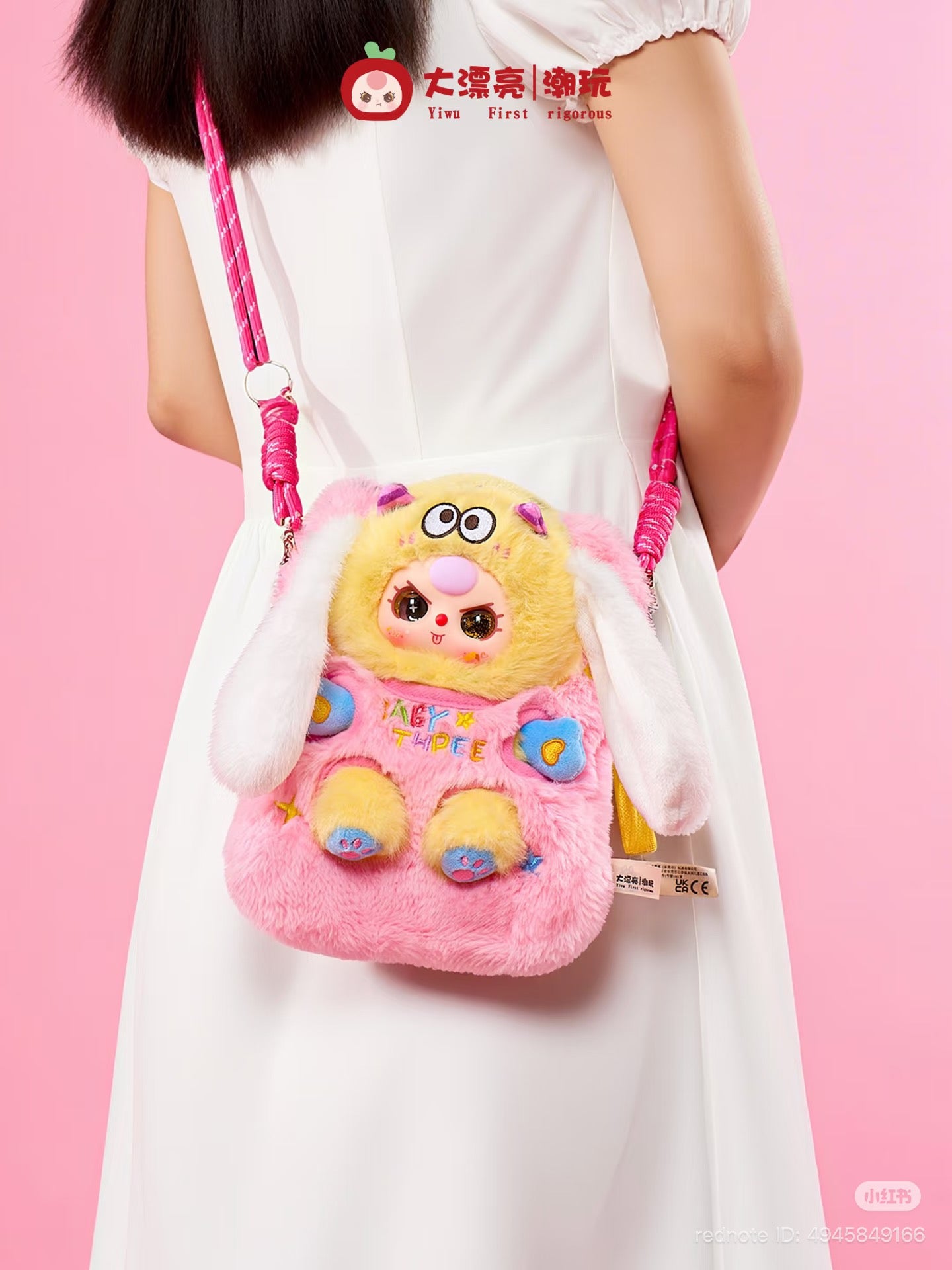 Person wearing Baby Three Plush Travel Bag, styled as a cartoon bunny with floppy ears, embroidered paws, and a pink rope strap against a pink backdrop.