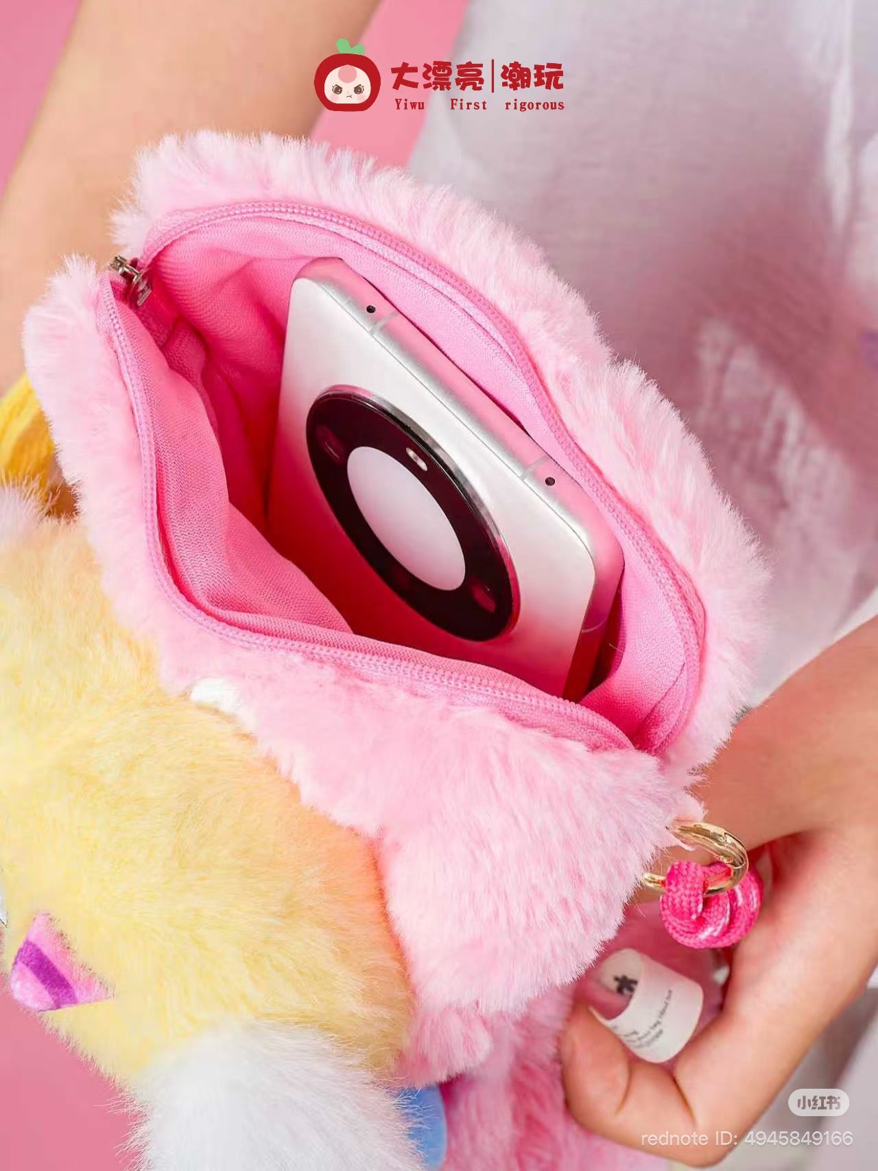 Baby Three Plush Travel Bag unzipped to show a silver gadget resembling a wireless charger. A hand holds the bag by its gold ring and pink strap.