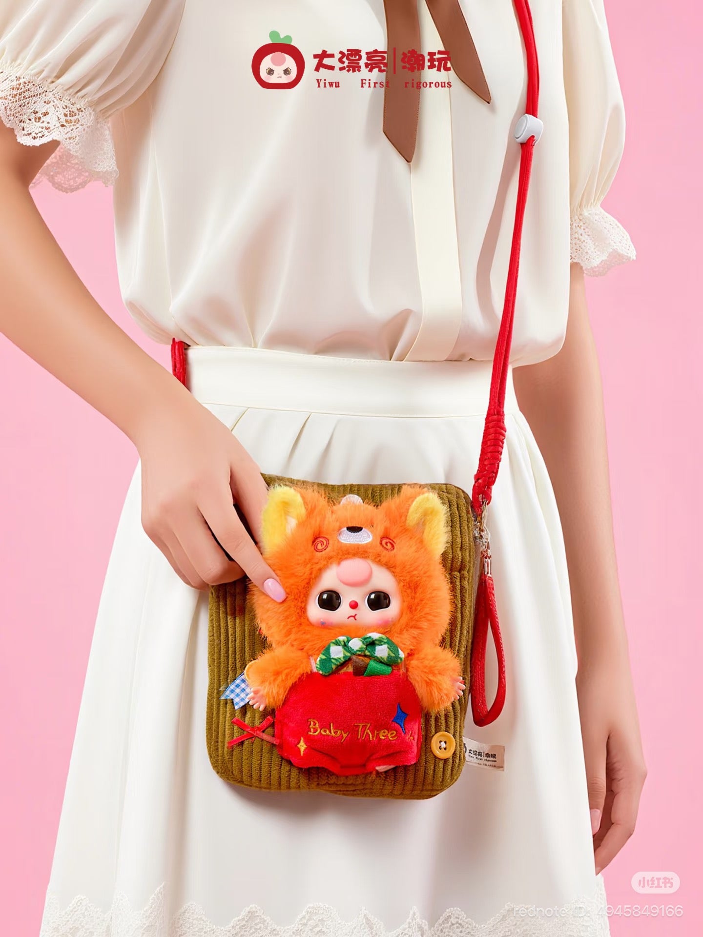 Model holds Baby Three Plush Travel Bag, featuring an olive corduroy design with a red strap and plush character in a hoodie on front.
