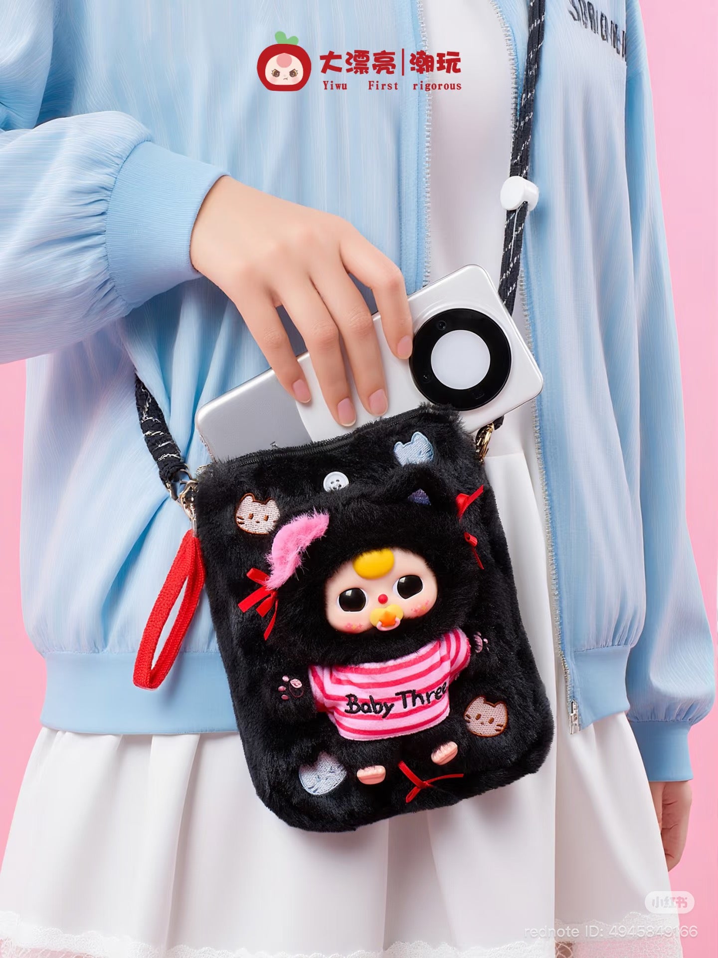Baby Three Plush Travel Bag with a baby-doll figure, embroidered cat faces, red bows, and detachable strap, shown with a phone being retrieved.
