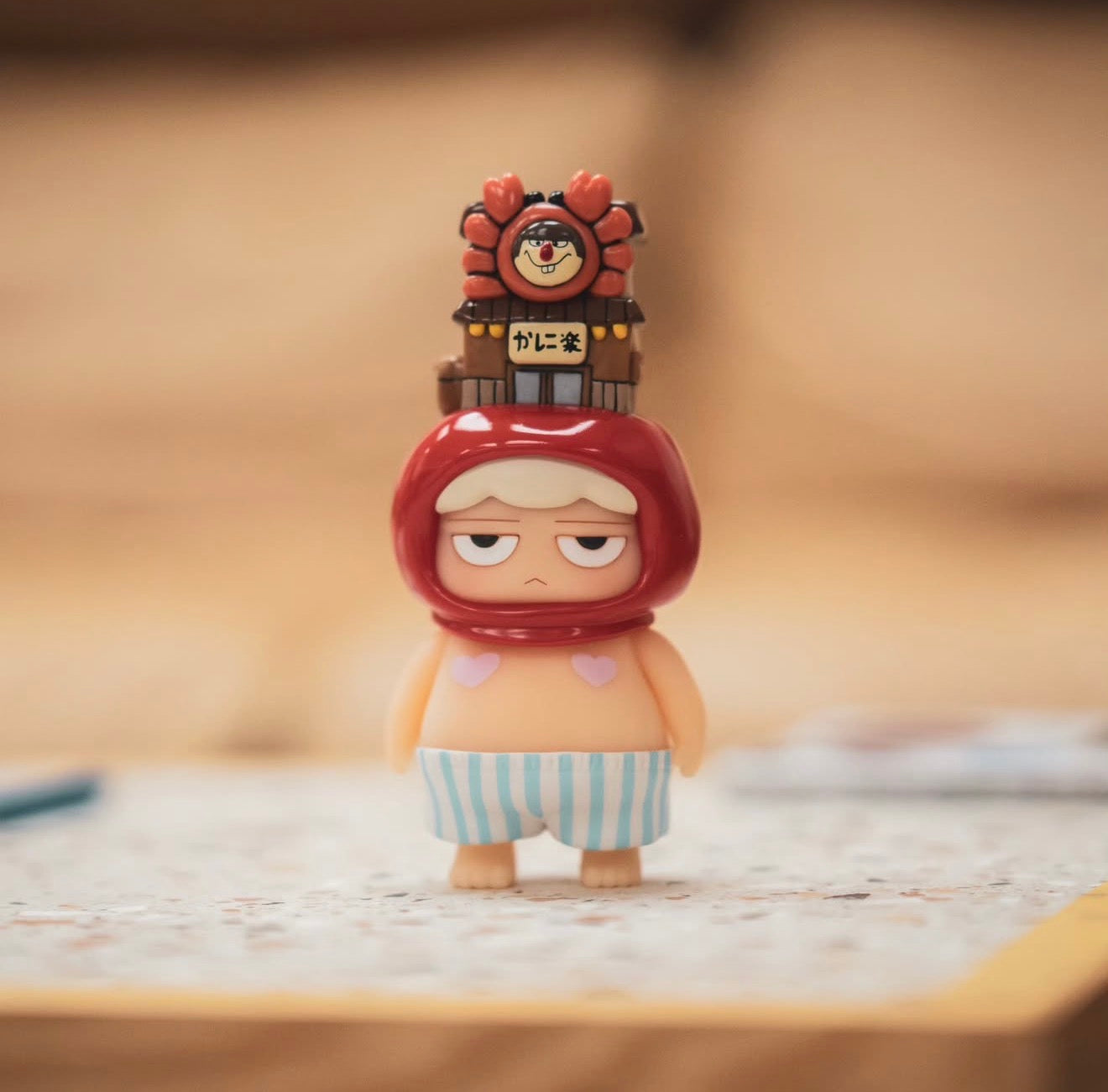 Poh x Worldsman vinyl figure: cartoonish character with red hood, shop façade on top, pink heart details, and striped shorts.