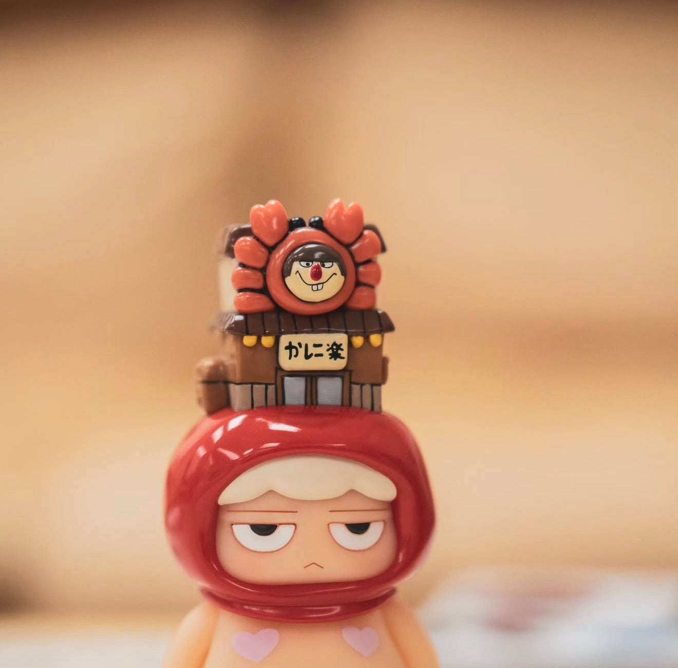 Poh x Worldsman figurine: a vinyl character in a red hood with droopy eyes, featuring a crab-themed shop on its head and heart decals.