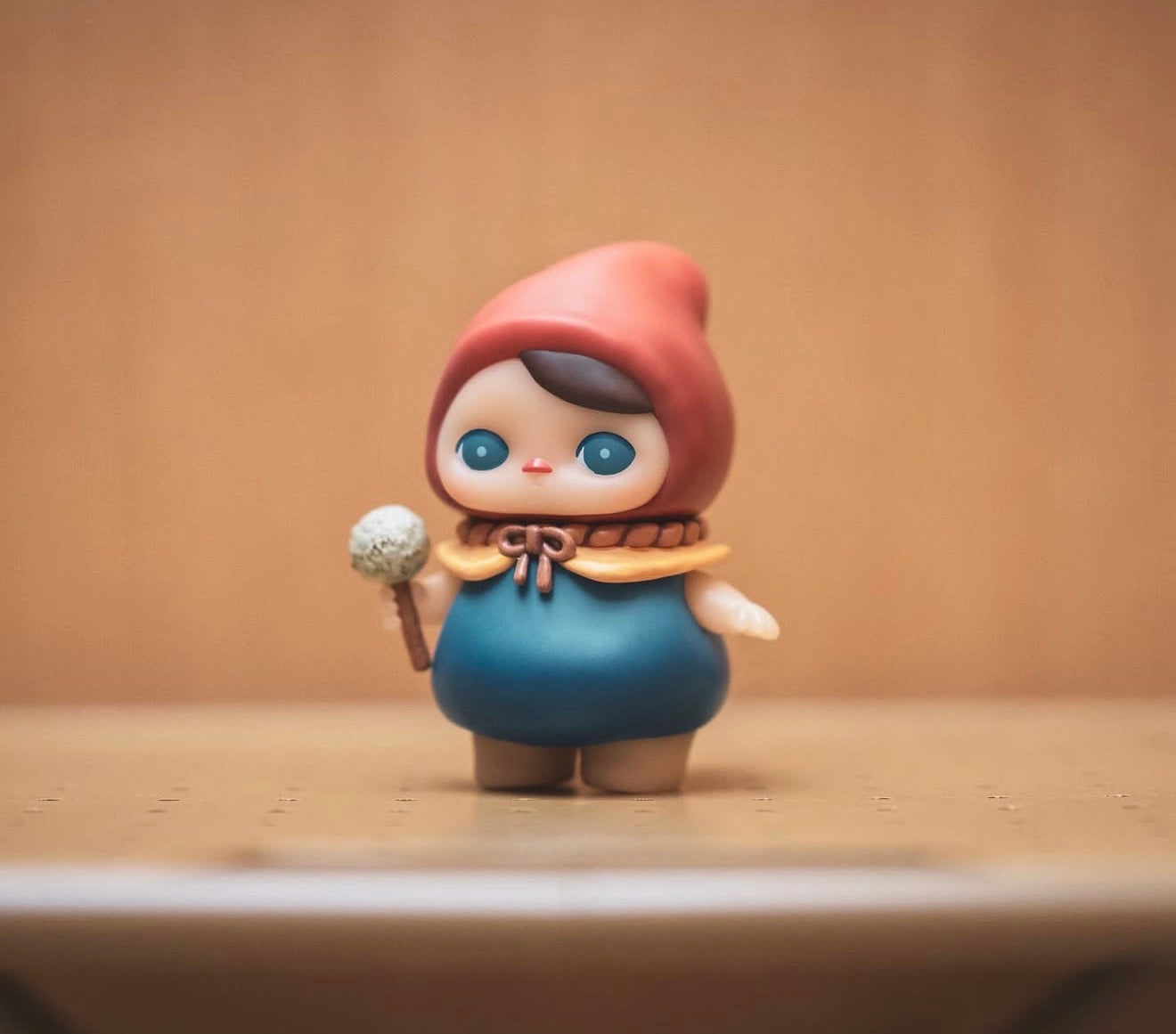 Pucky Poko vinyl figurine in a red hood, yellow cape, and blue tunic, holding a tiny flower, showcased on a wooden surface.