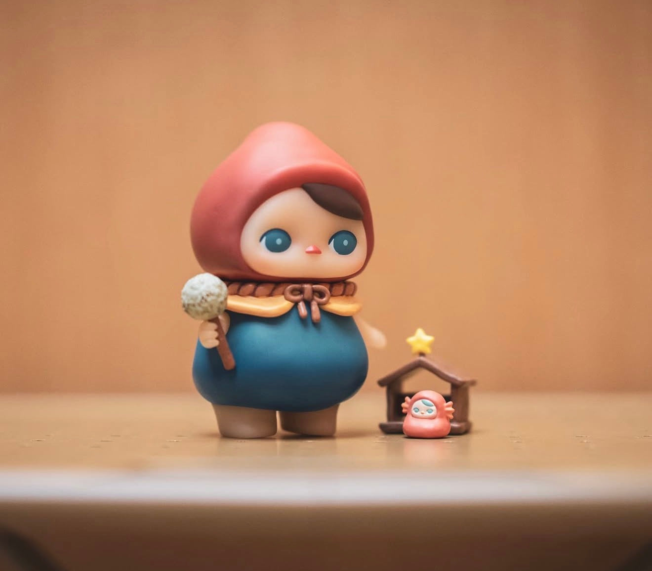Pucky Poko vinyl toy figure holding a dandelion puff, set beside a tiny hooded figure and stable scene, evoking a whimsical, storybook charm.