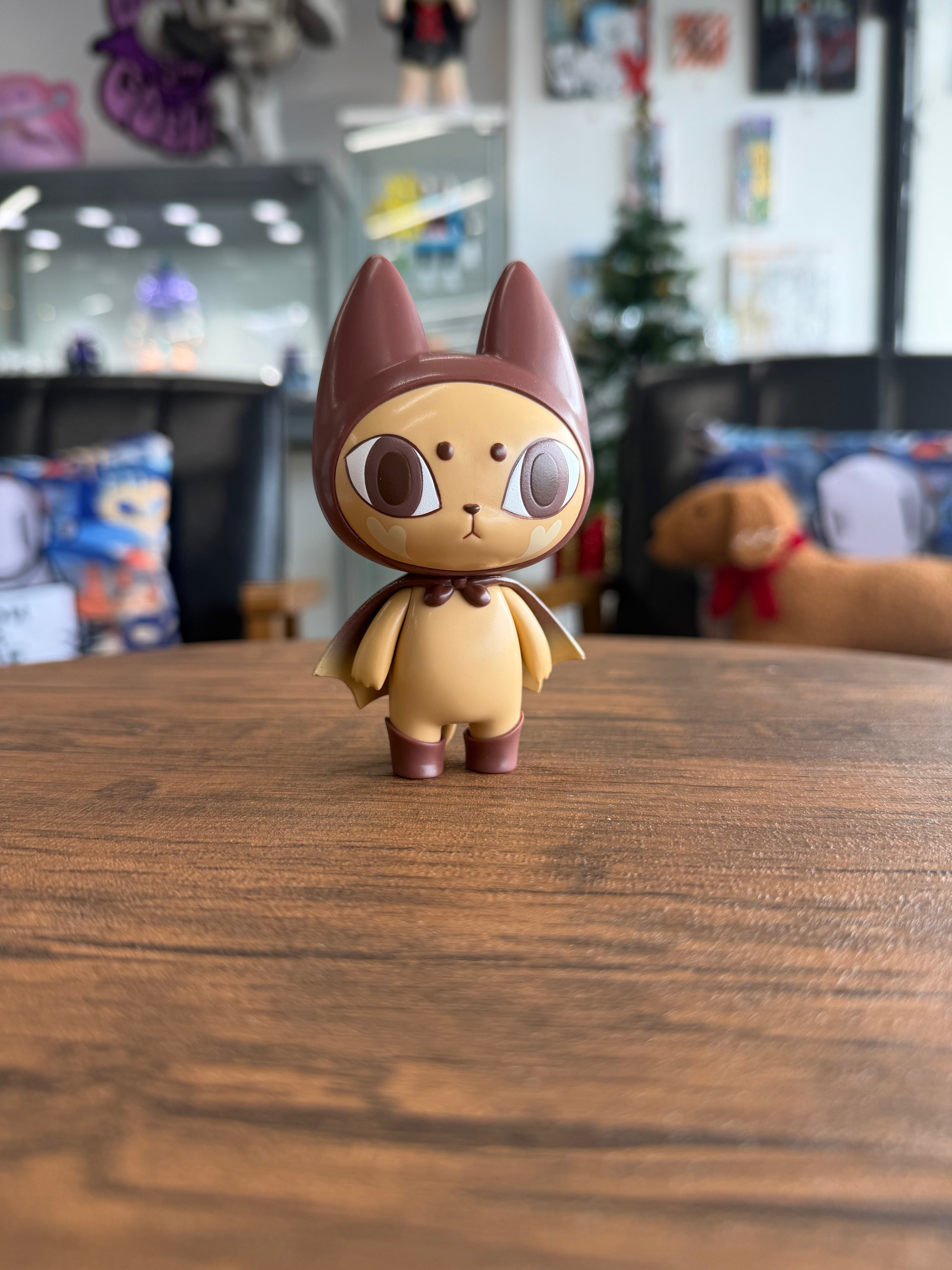BAD MEAW 'Latte' EDITION by Mueanfun Sapanake: a 4 vinyl toy cat in bat-inspired attire, displayed on a wooden tabletop amidst collectibles.