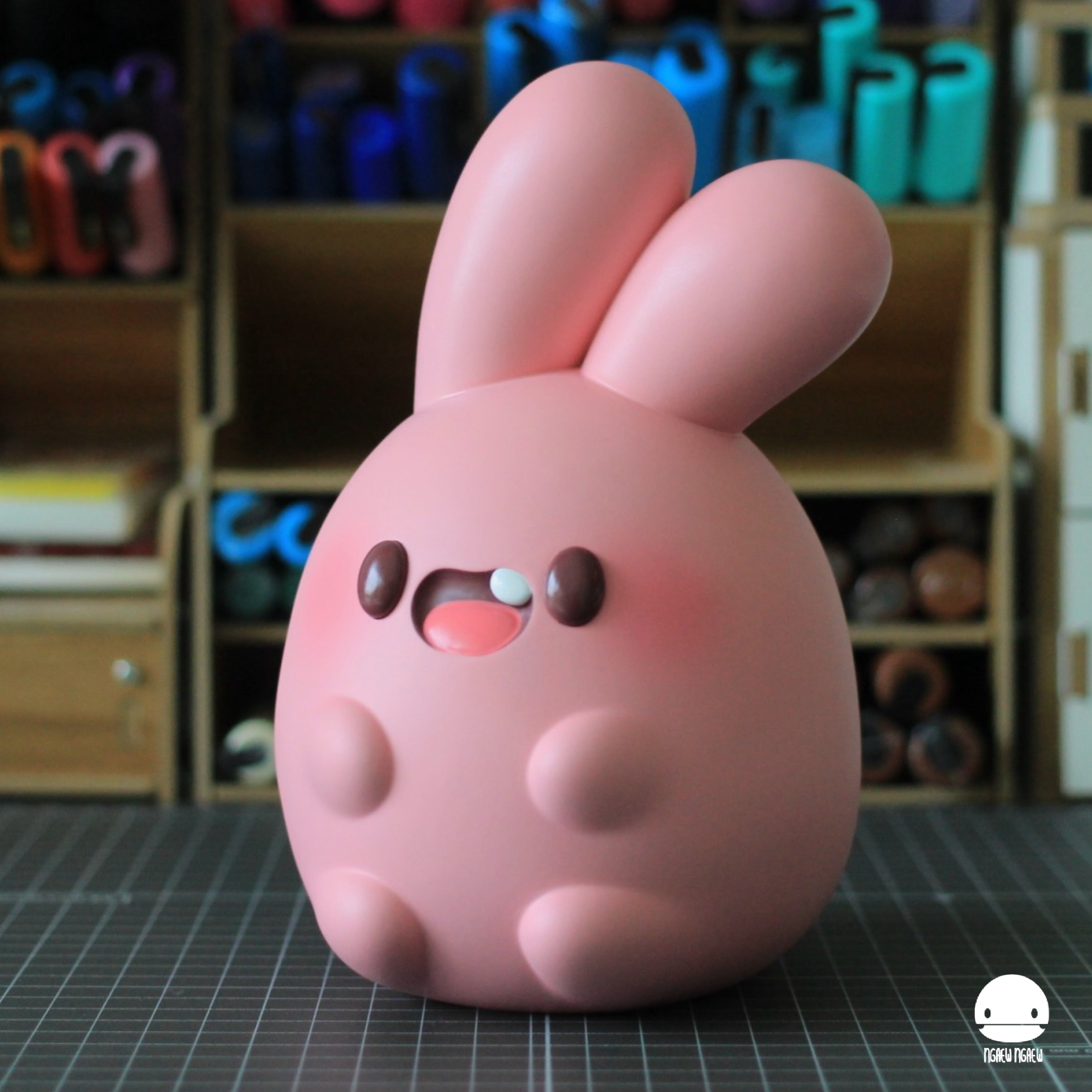 NGAEW Heavy by Ngaew Ngaew: A pastel-pink, chubby bunny figurine with long ears and a smiling mouth, sitting on a cutting mat, 18cm resin, 3lbs.