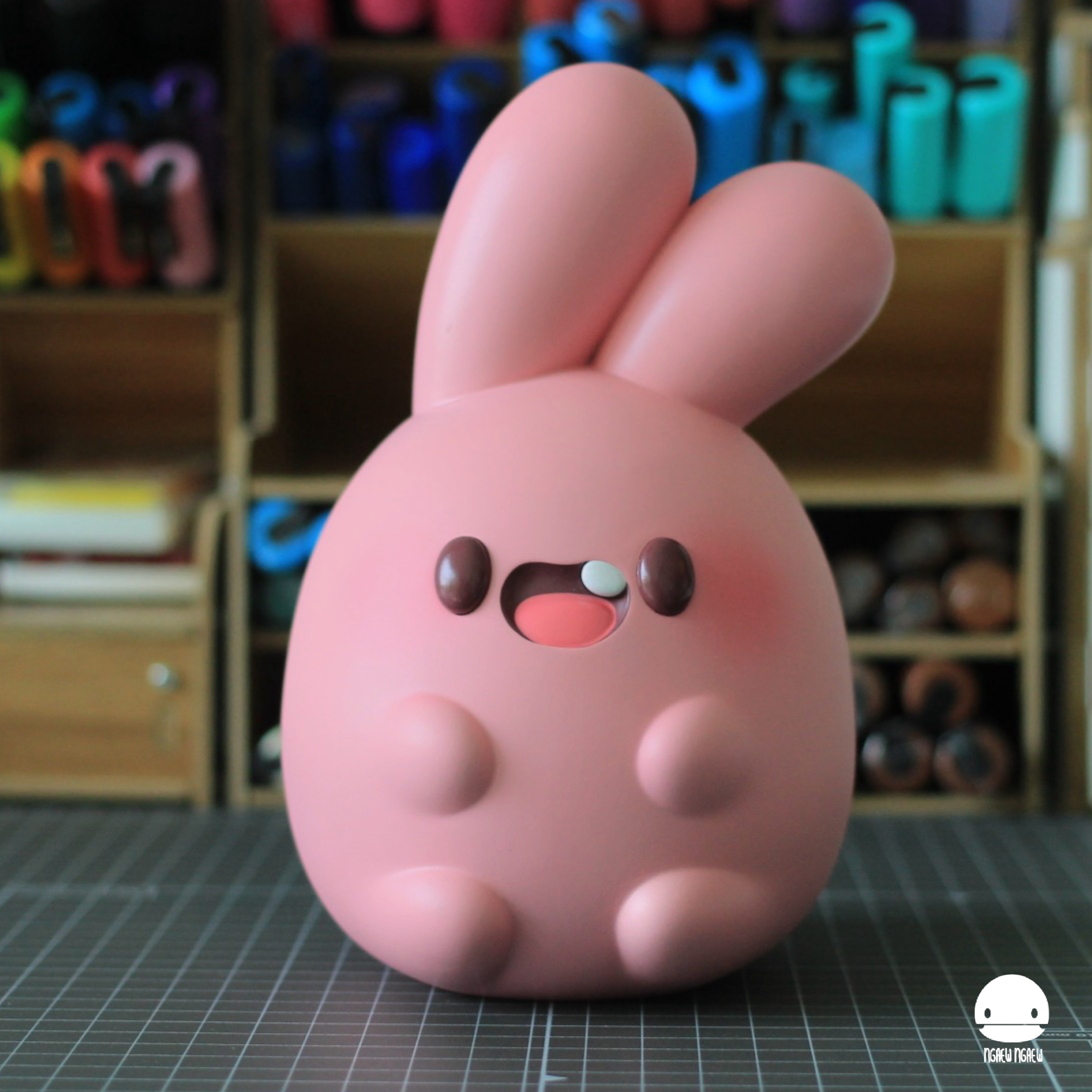 NGAEW Heavy by Ngaew Ngaew: A pink, stylized vinyl bunny figure with an egg-shaped body, long ears, and a happy face, set on a cutting mat.