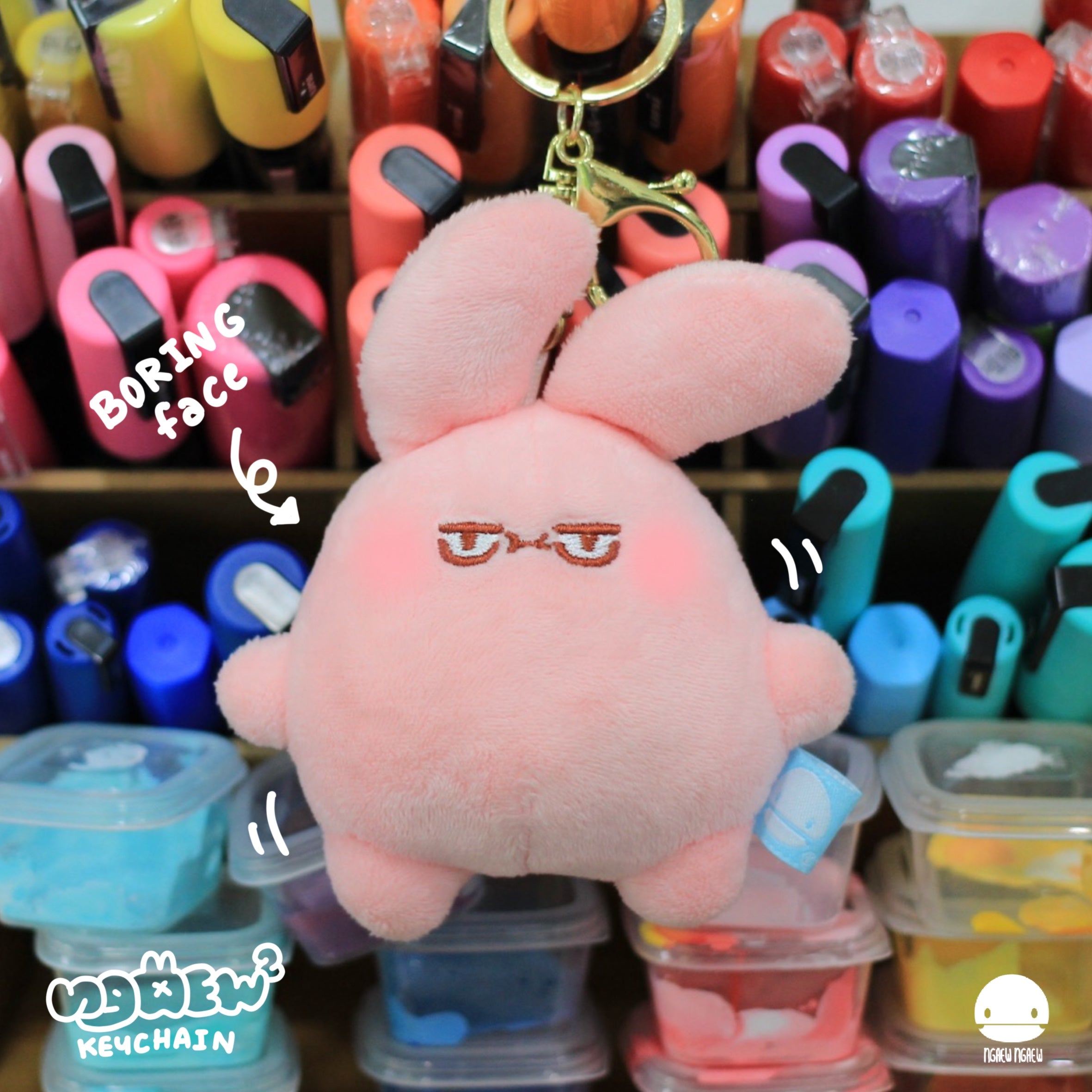 Ngaew Ngaew Plush Keychain: pink rabbit with floppy ears, bored expression, gold clip. Background features colorful items, enhancing the playful, artistic vibe of Strangecat Toys.