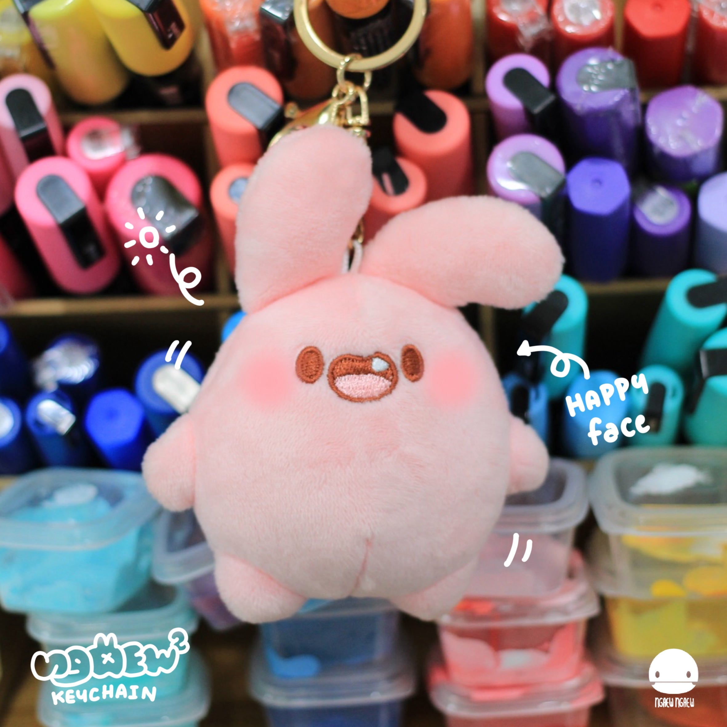 Ngaew Ngaew Plush Keychain, a small pink bunny with floppy ears and a smiling face, surrounded by art supplies and playful doodles.