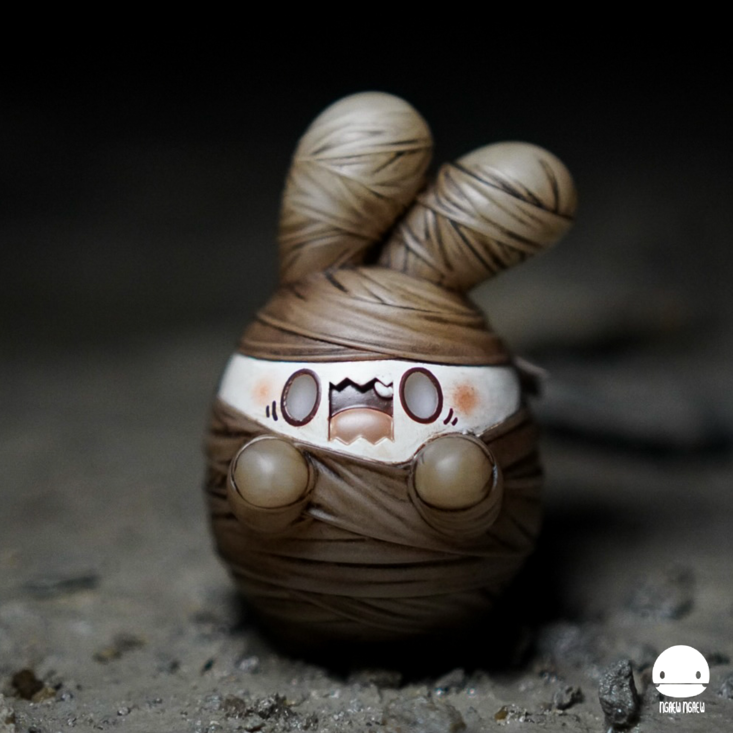 Ngaew Ngaew Mummy figurine; a resin bunny-like creature wrapped in bandages, with exposed face details, on a dirt surface. Limited edition, 8 cm tall.