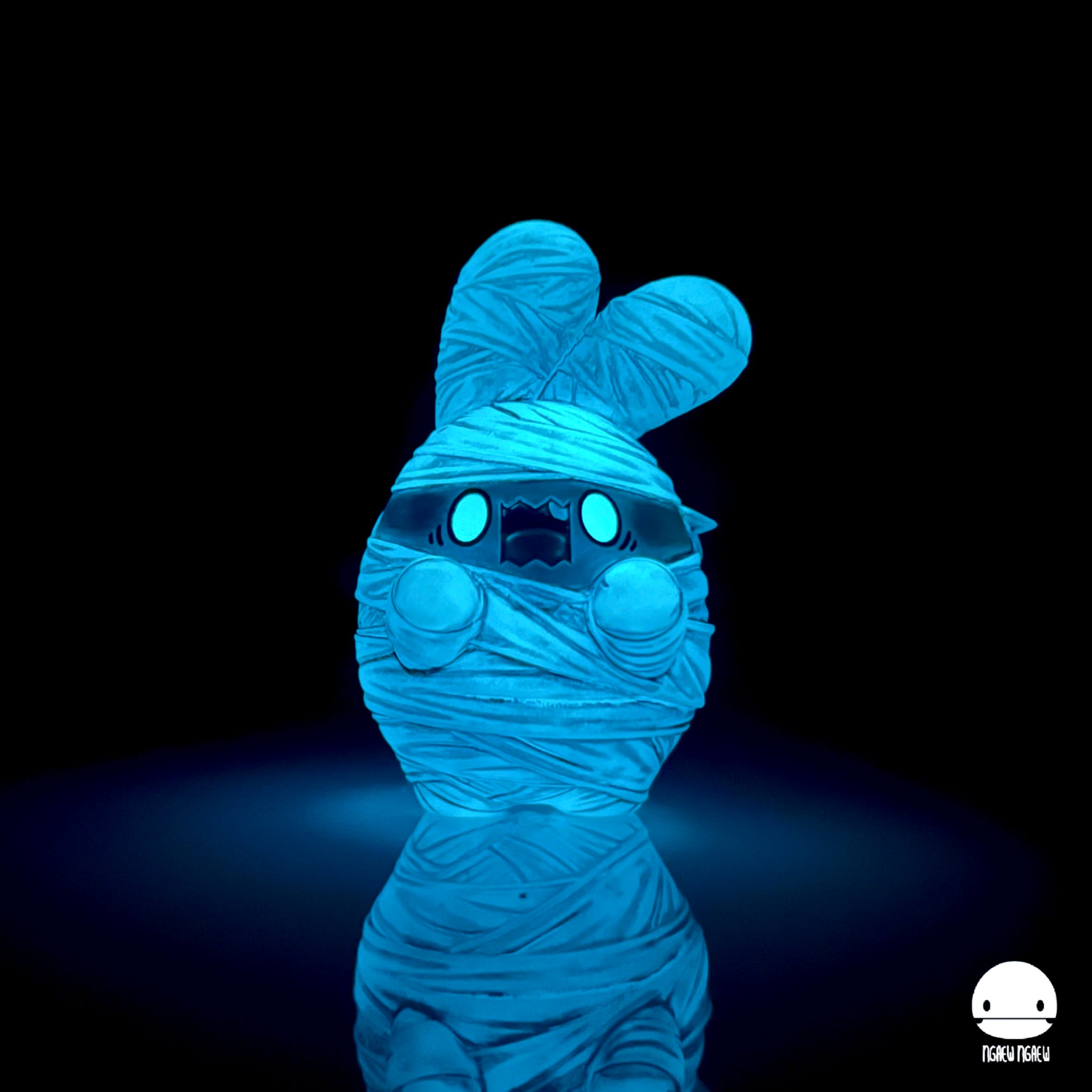 Ngaew Ngaew Mummy by Ngaew: a glowing resin figure with bandages, bunny ears, and blue eyes, standing on a reflective surface. Limited edition, 8 cm.