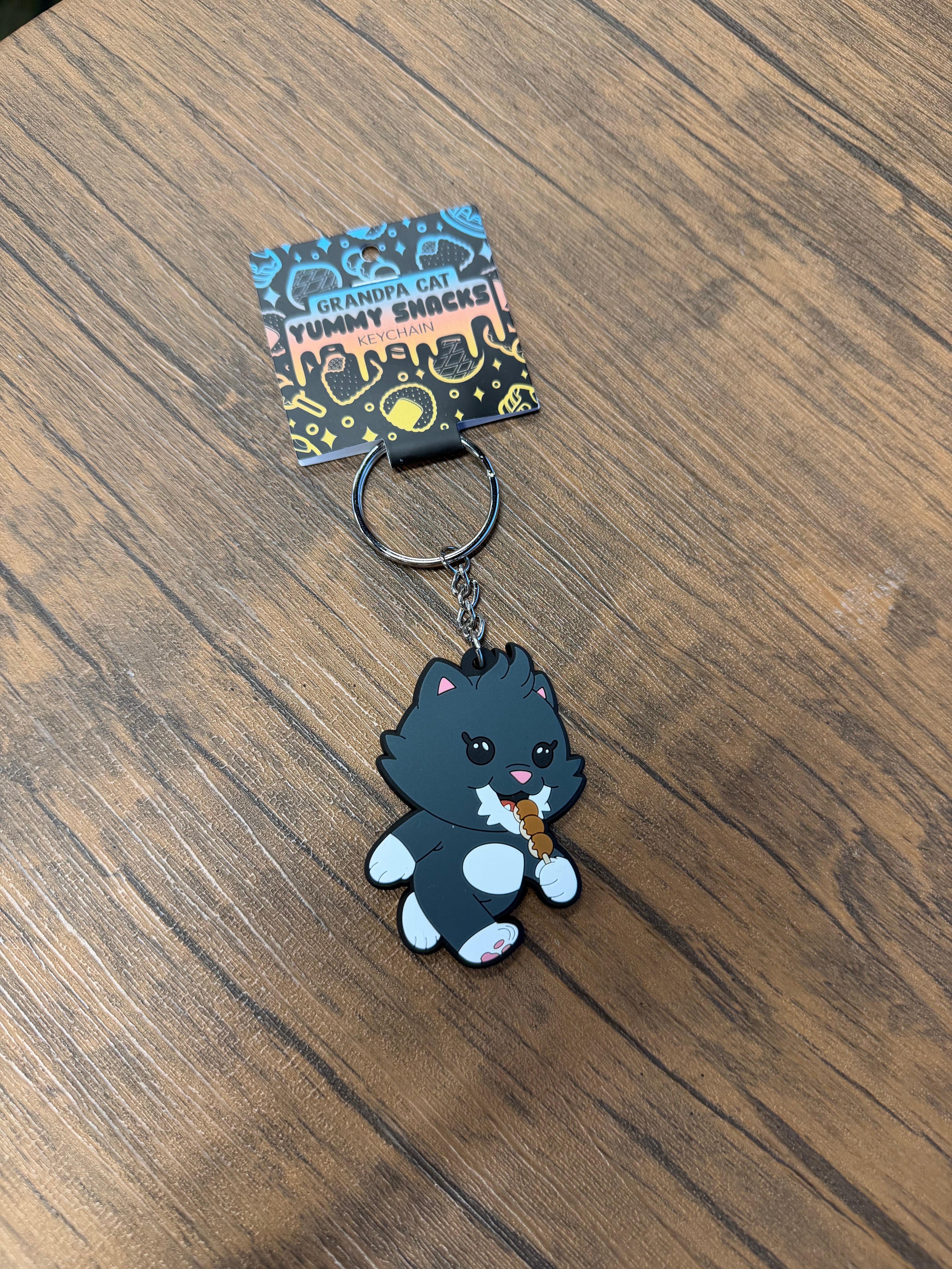 Grandpa Keychain - Japanese Snack Series: A cartoon-style cat figure nibbling snacks, attached to a keyring, displayed on a wood-grain surface. Silicone, 2.75 inches.