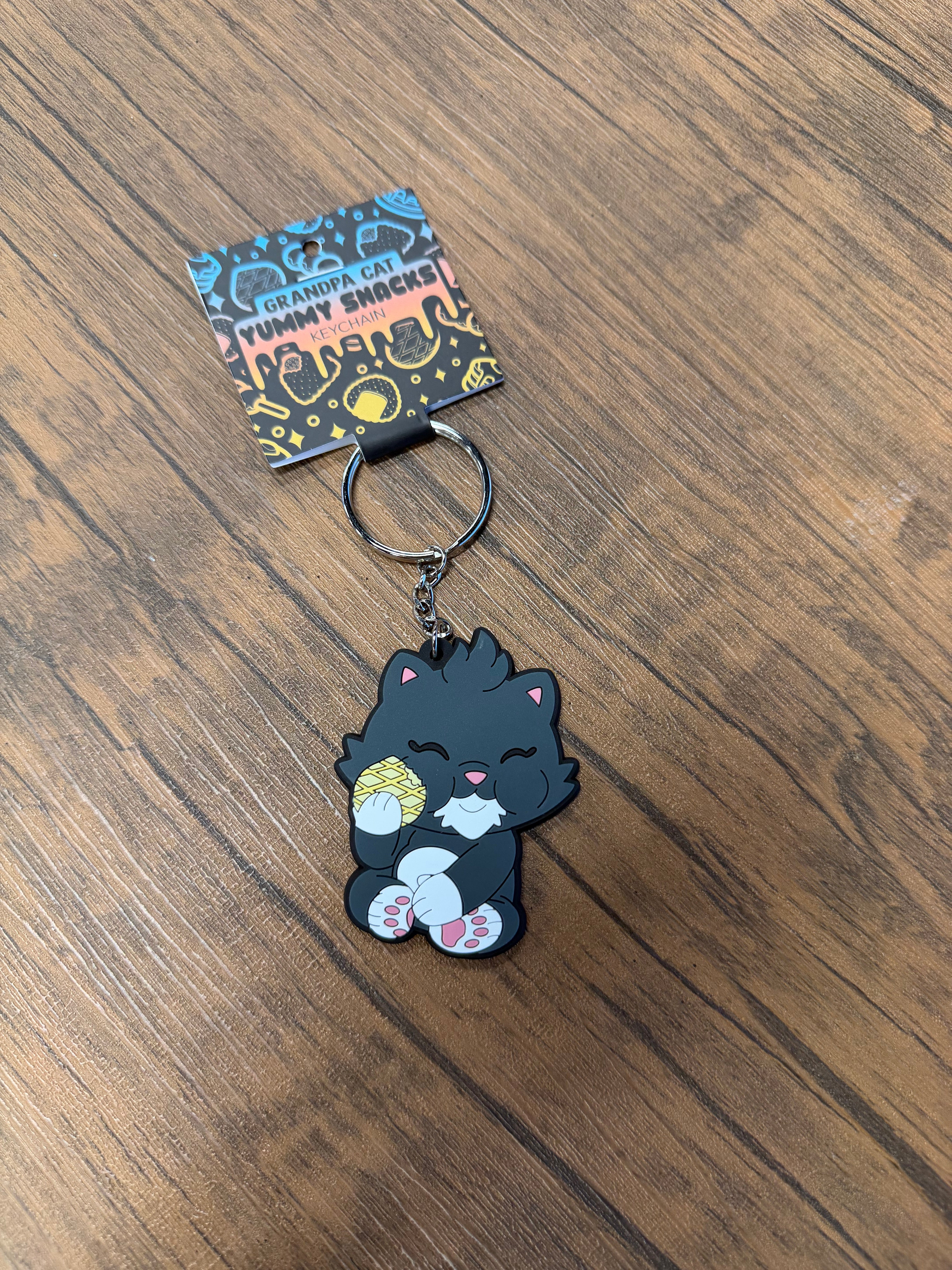 Grandpa Keychain - Japanese Snack Series features a rubber charm of a cat holding a pastry, displayed on a branded card atop a wooden surface.