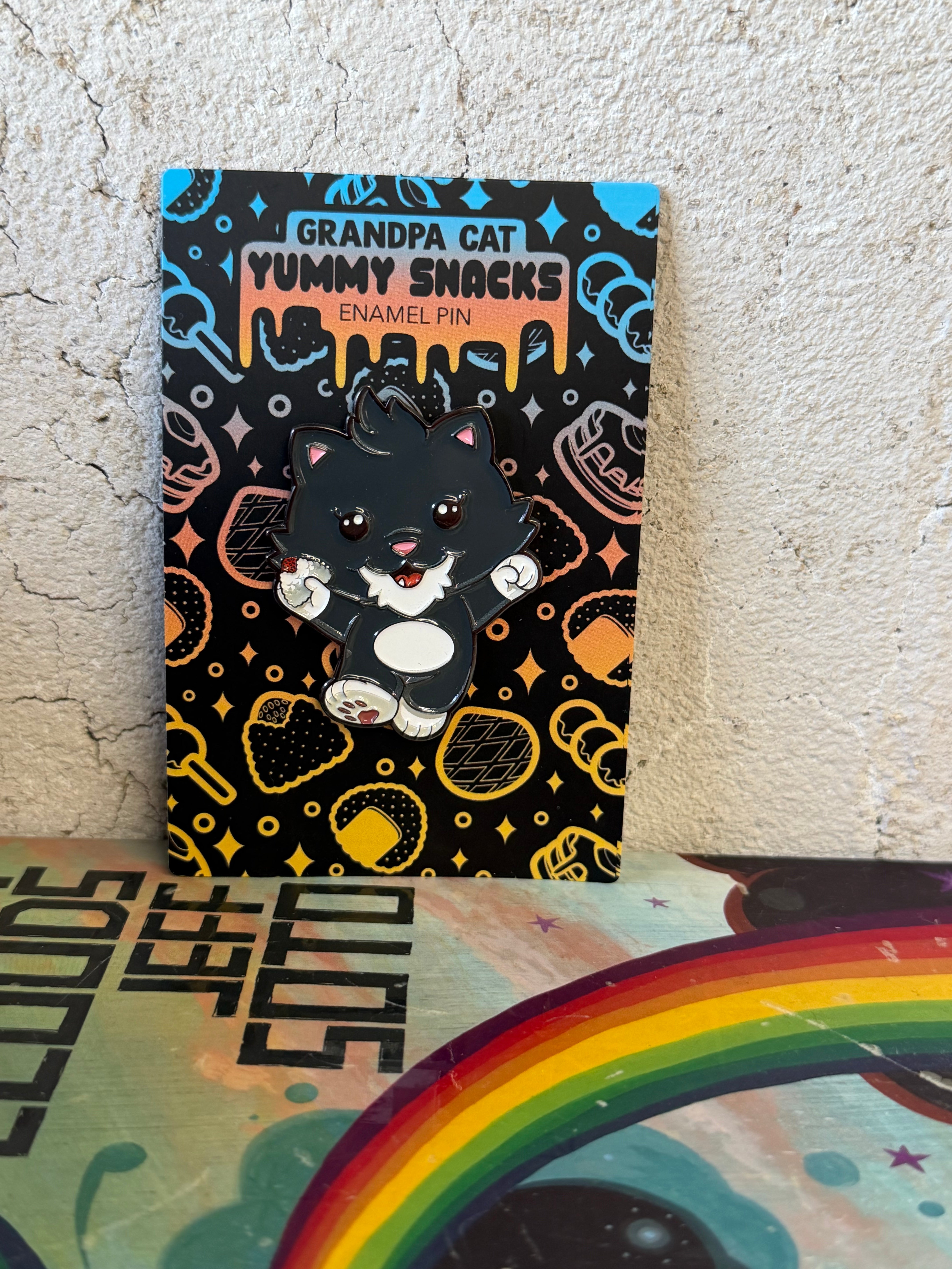 Grandpa Pin - Japanese Snack Series: A 2 enamel pin of a cartoonish black-and-white cat holding a treat, set against a snack-themed backing card.