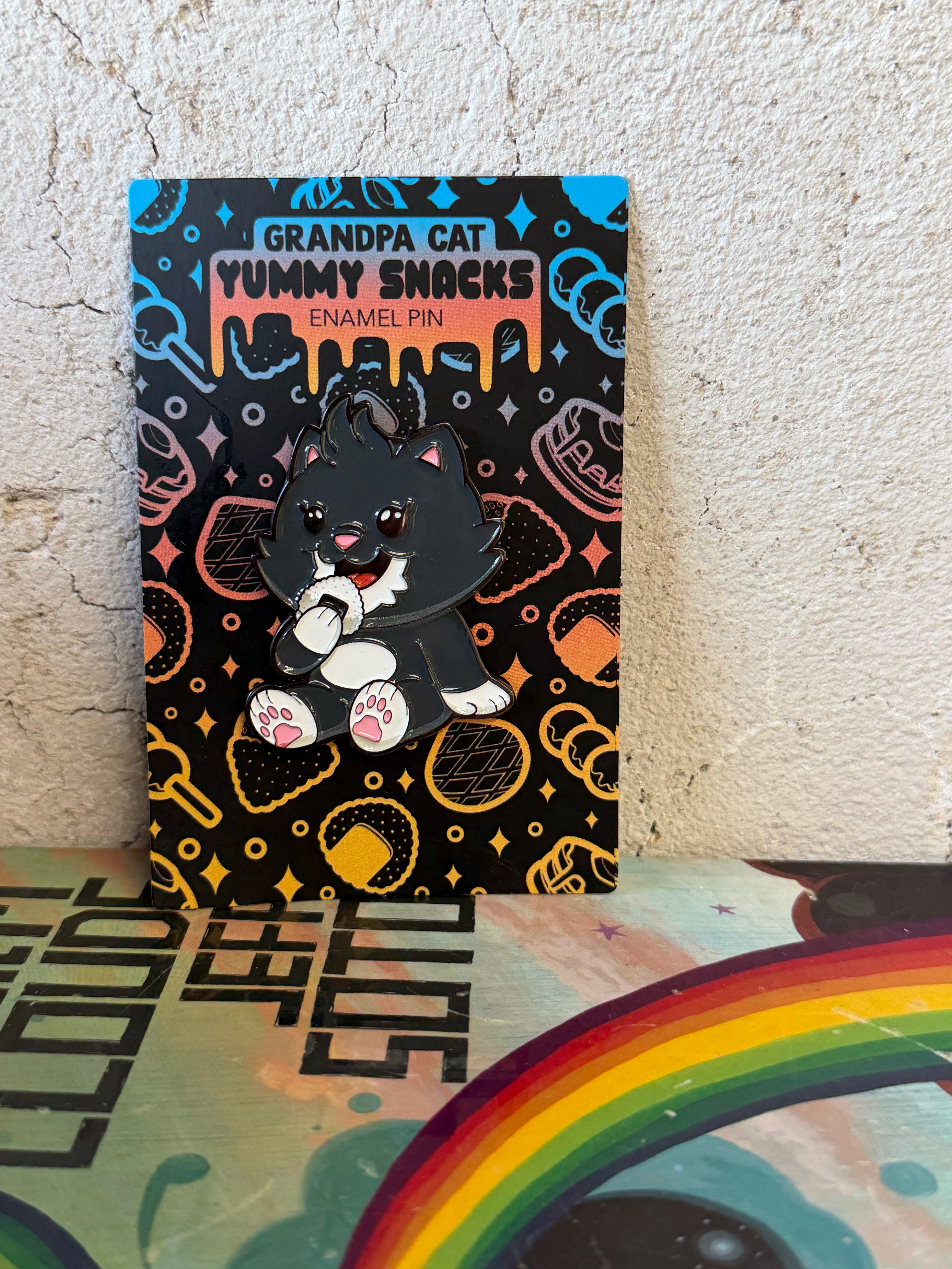 Grandpa Pin - Japanese Snack Series: A cartoon black cat enamel pin from Strangecat Toys, depicted nibbling a cookie, showcased on a colorful, snack-themed backing card.