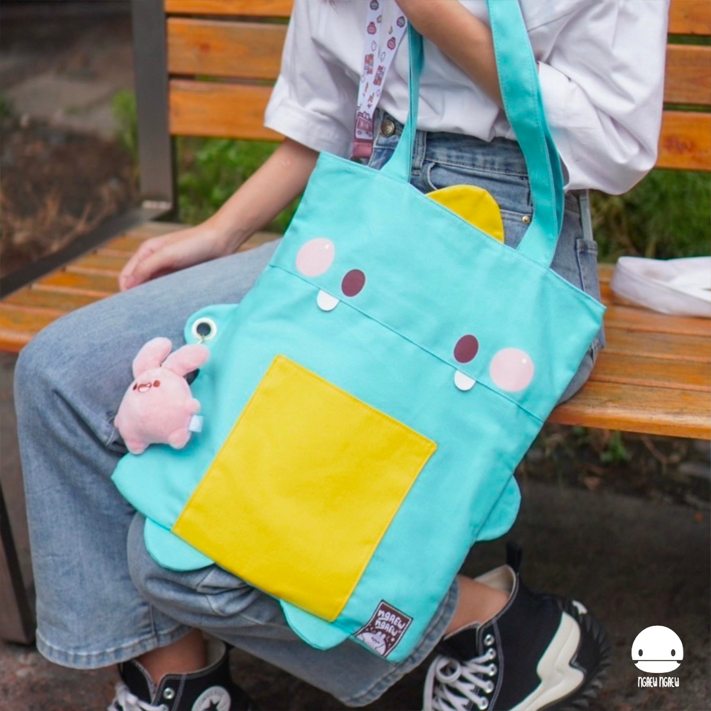 Person holding NGAEW NGAEW TOTE BAG, an aqua canvas tote with cartoon features, yellow pocket, and pink bunny keychain, reflecting Strangecat Toys' playful style.