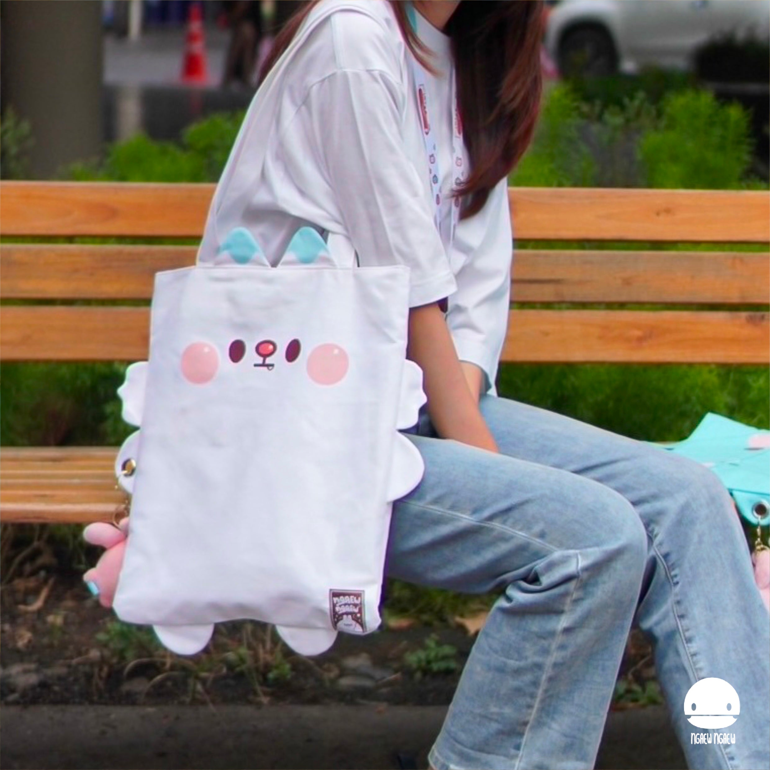 Person on a bench with NGAEW NGAEW TOTE BAG, featuring cartoon creature design with floppy ears and rosy cheeks. A turquoise bag with plush keychain is partially visible.