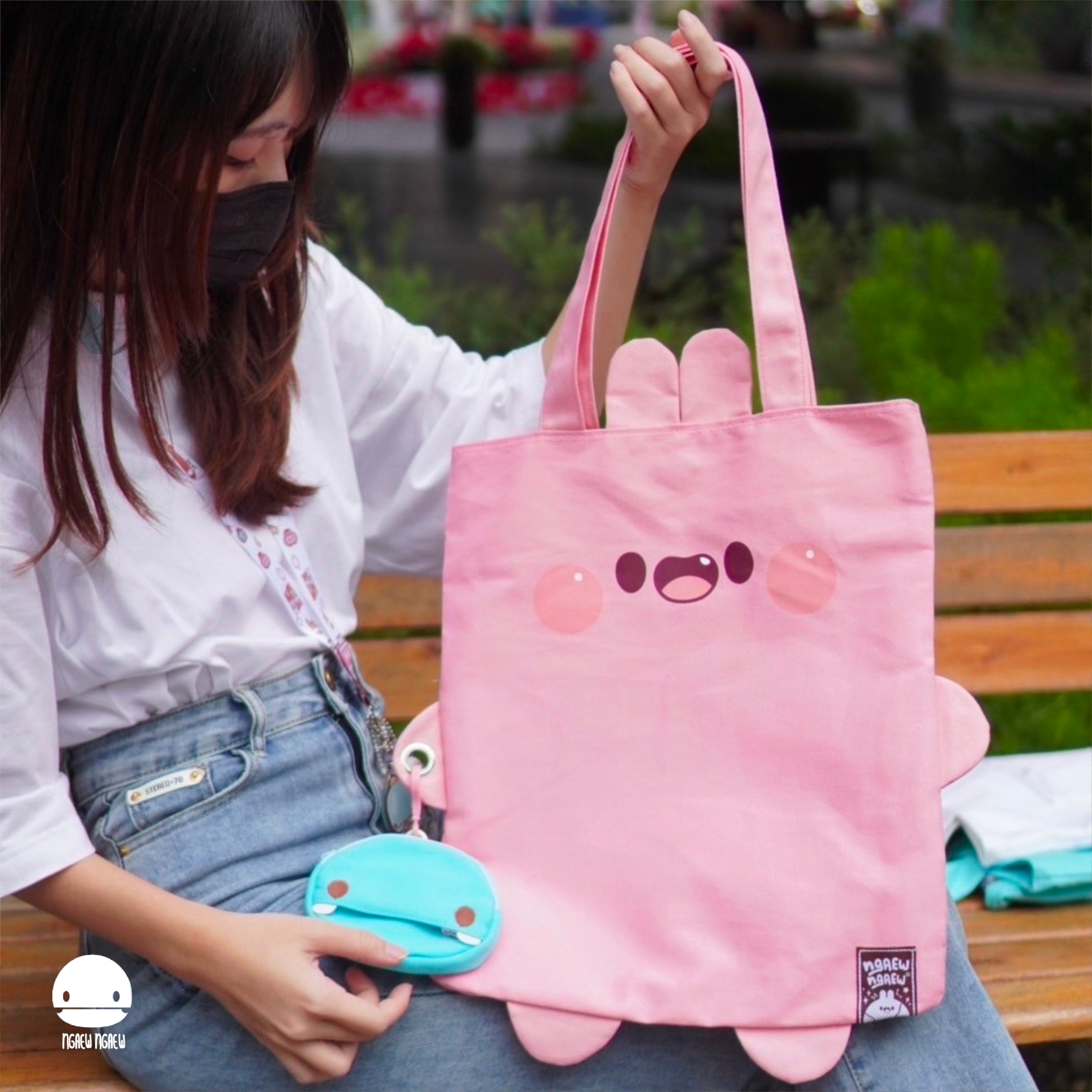 A person displays the NGAEW NGAEW TOTE BAG, featuring a kawaii face and ear tabs, with a blue snap-pouch on the handle.