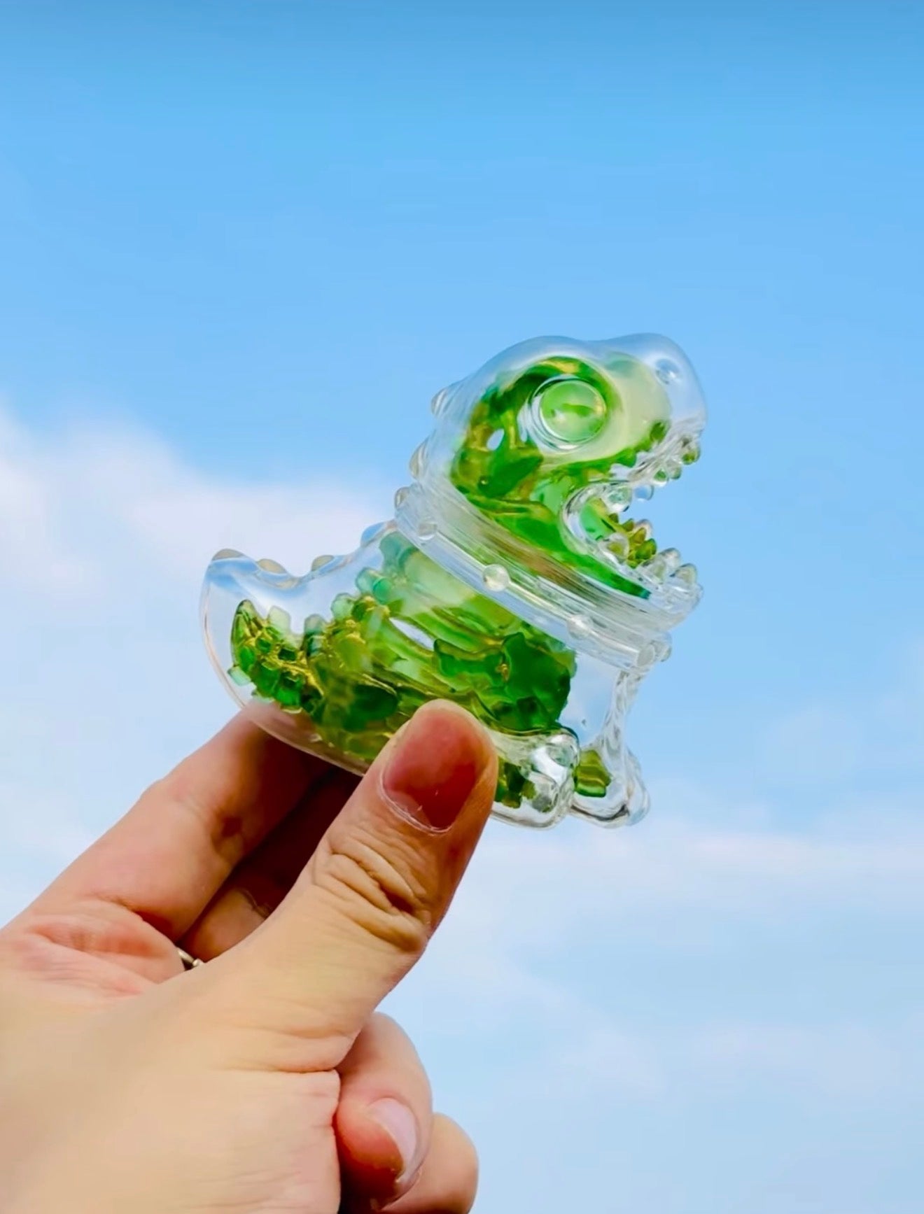Person holding the Emerald Skeleton Dragon by HOTRU, a clear plastic dinosaur with green gel, against a blue sky. Preorder for playful toy enthusiasts.
