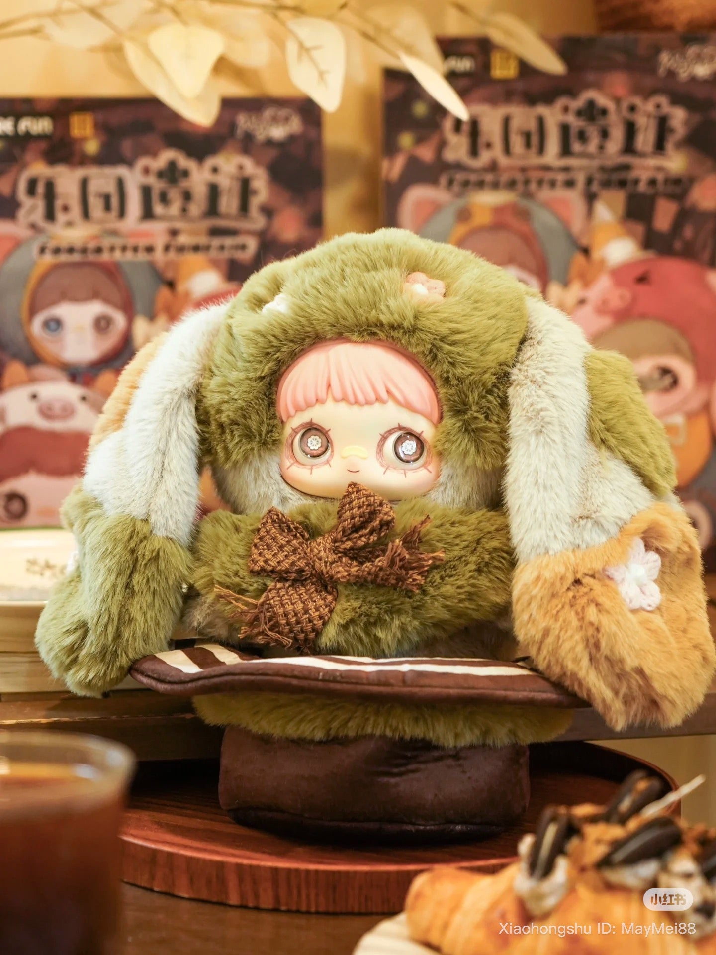Maymei 400% Forgotten Funfair Plush Keychain Blind Box figure in green bunny suit, seated with coffee and pastry, colorful packaging blurred behind.