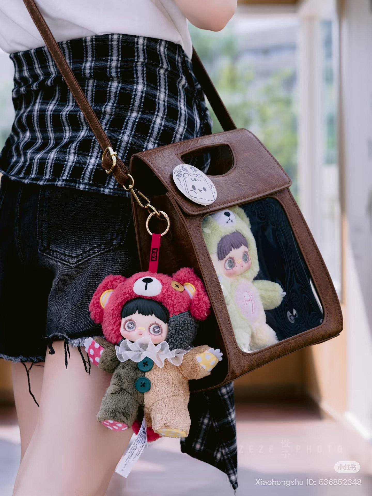 Close-up of black shorts, plaid shirt, and a small brown cross-body bag with MayMei Symbiote Neverland Series plush toys and keychain.