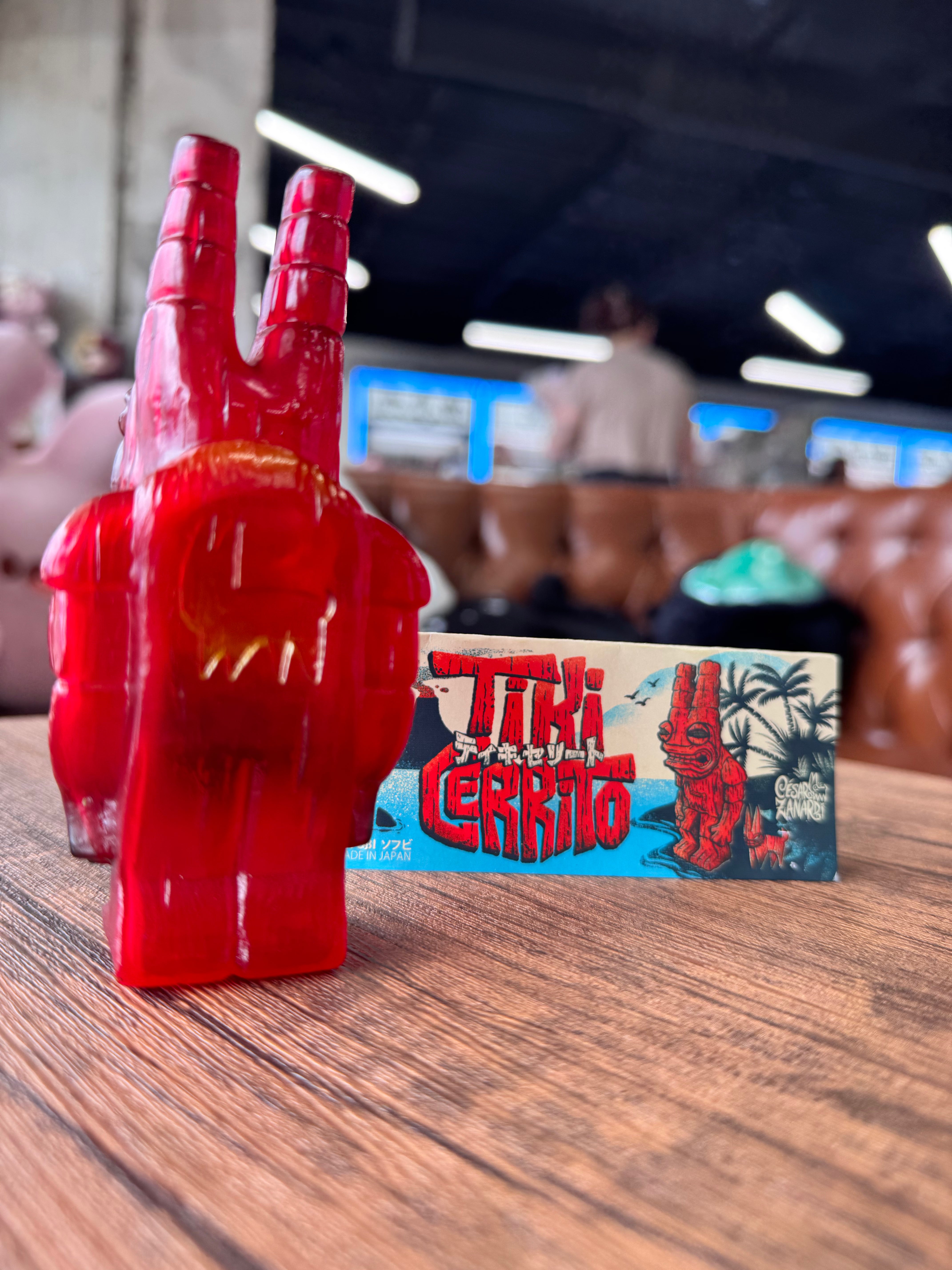 Tiki Cerrito by Cesar Zanardi, a 12cm red vinyl toy with horn-like features, stands on a tabletop with its acrylic background featuring tiki artwork.