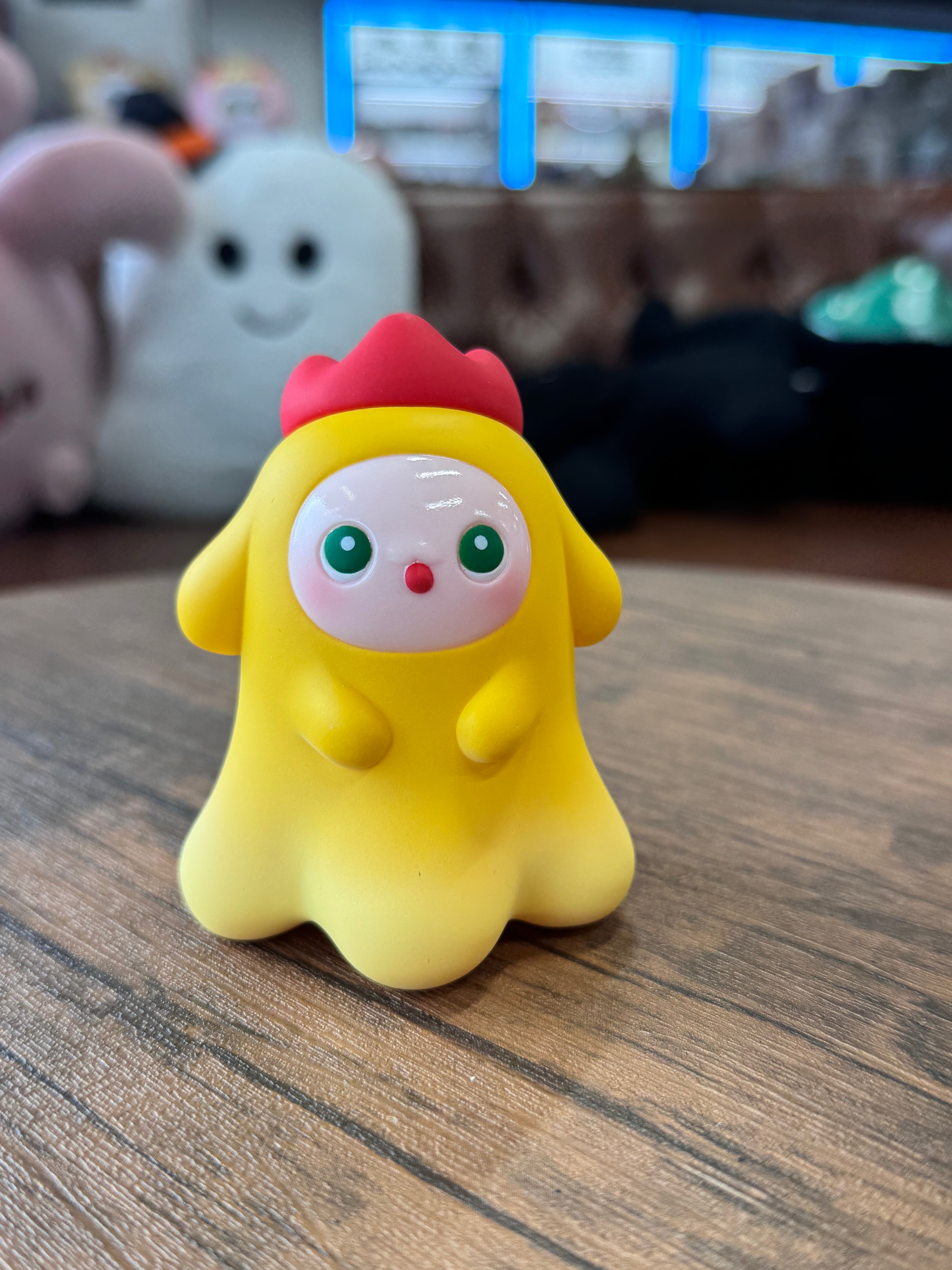 MYONNY - yellow by PYOS: A 9cm ghost-shaped toy figure with a red crest, large green eyes, and rosy cheeks, seated on a wooden tabletop.