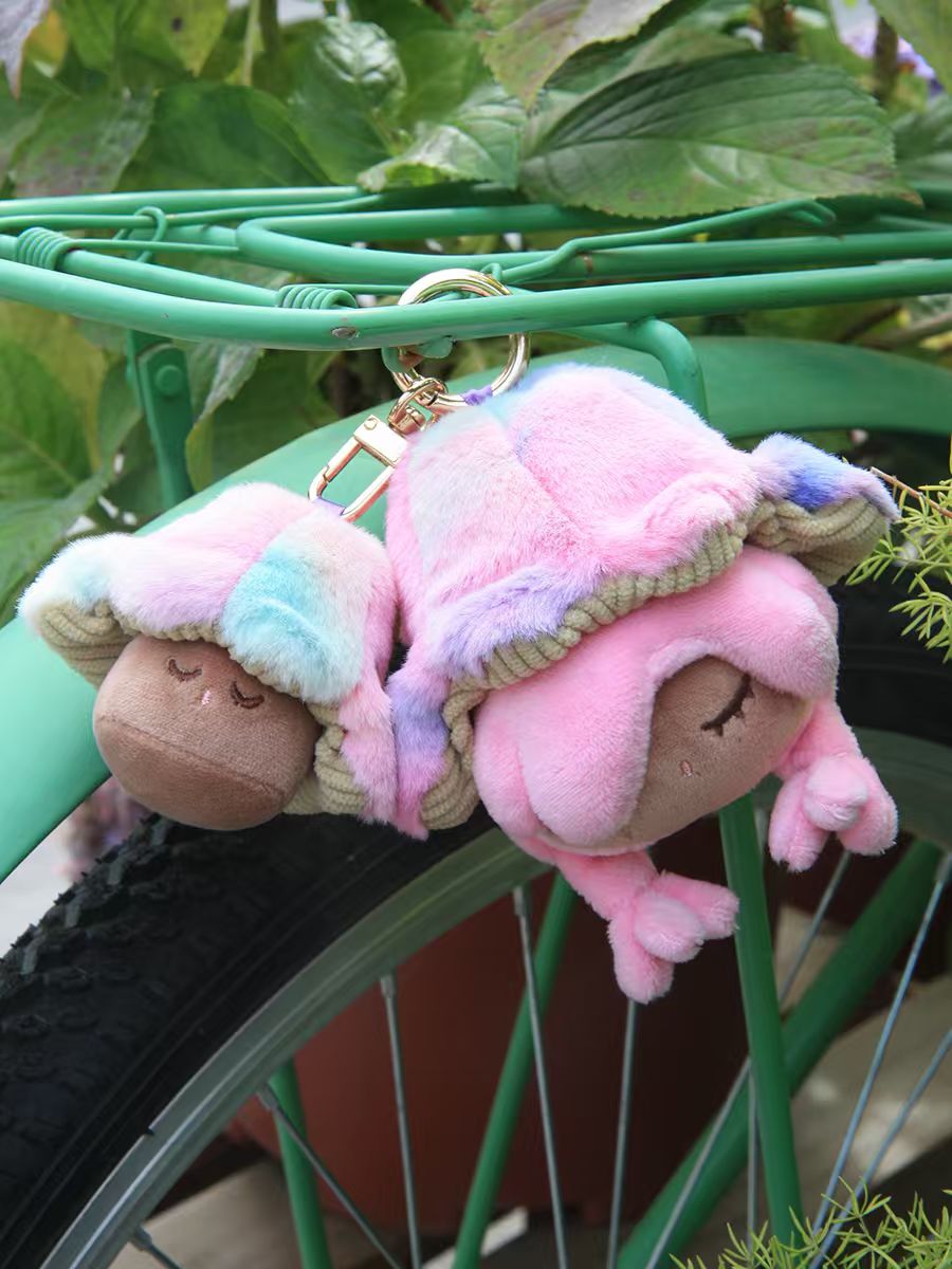 Little Mushroom Plush Pendant Blind Box keychains, featuring sleepy faces and floppy limbs, dangle from a bike rack, showcasing the whimsical charm of Strangecat Toys.